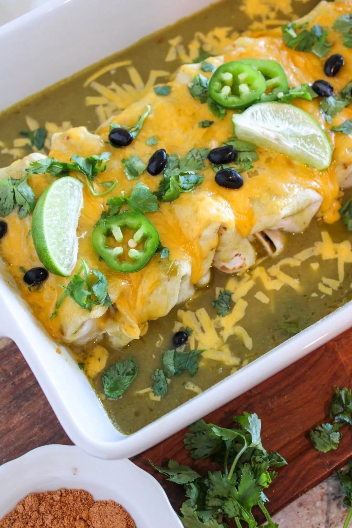 Cheesy enchiladas in green sauce, topped with lime wedges, sliced jalapeños, black beans, and cilantro; a colorful, appetizing dish.