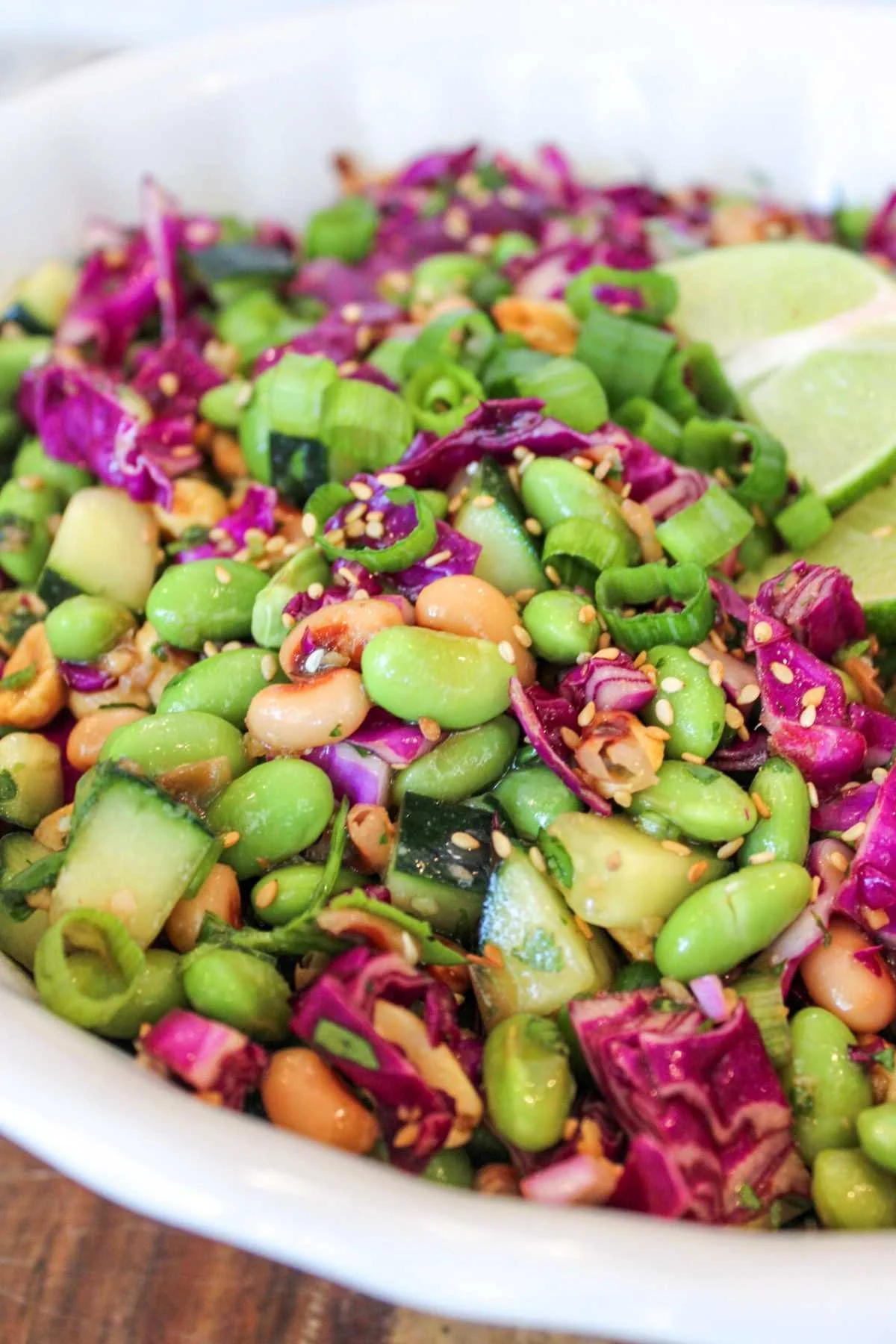 Fresh edamame bean salad with vibrant purple cabbage, cucumbers, peanuts, and sesame seeds in a white bowl, garnished with lime slices and green onions.