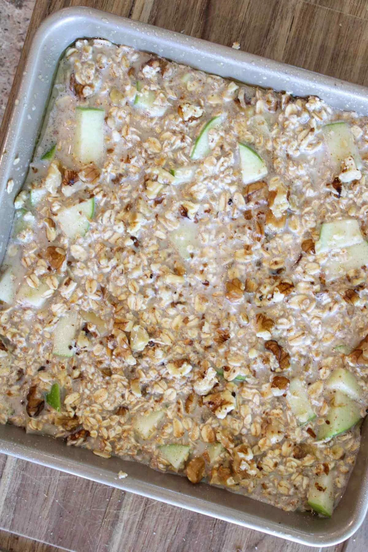 A square metal baking pan holds a rustic apple-oat mixture with rolled oats, diced green apples, cinnamon, and a moist, textured surface.