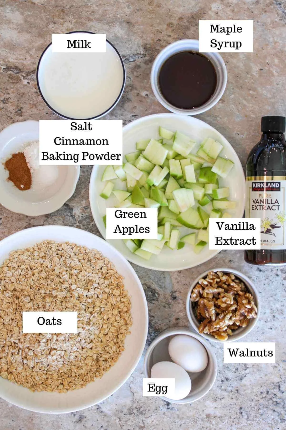 Overhead view of baking ingredients on a stone counter: oats, green apples, milk, egg, walnuts, maple syrup, vanilla, salt, cinnamon, and baking powder.