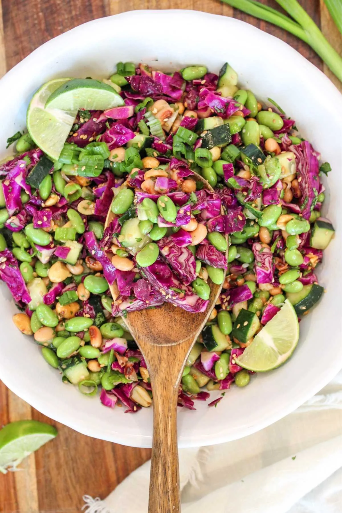 A vibrant salad in a white bowl with red cabbage, edamame, cucumber, and peanuts. A wooden spoon and lime wedges add freshness and texture.