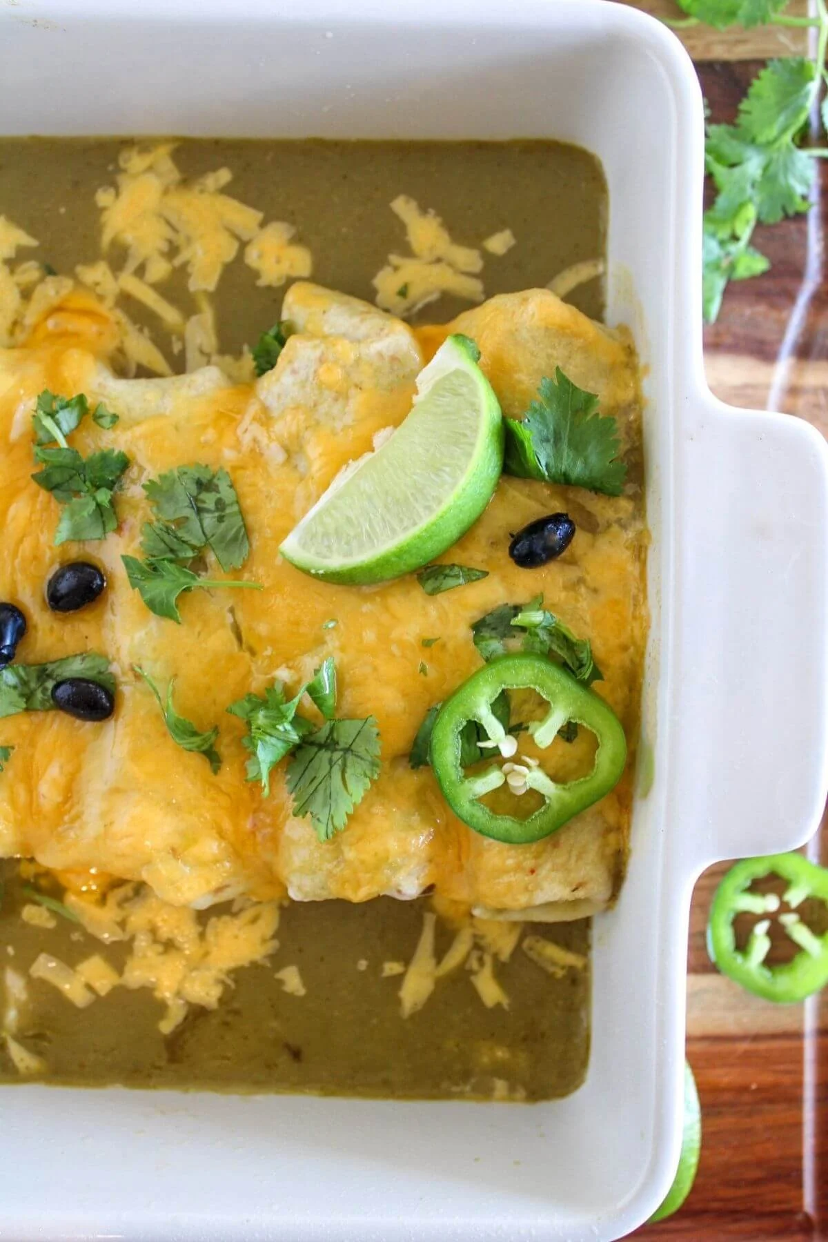 Golden baked chicken and black bean enchiladas in green sauce, topped with lime, cilantro, jalapeño slices, melted cheese, and black beans in a white casserole dish.