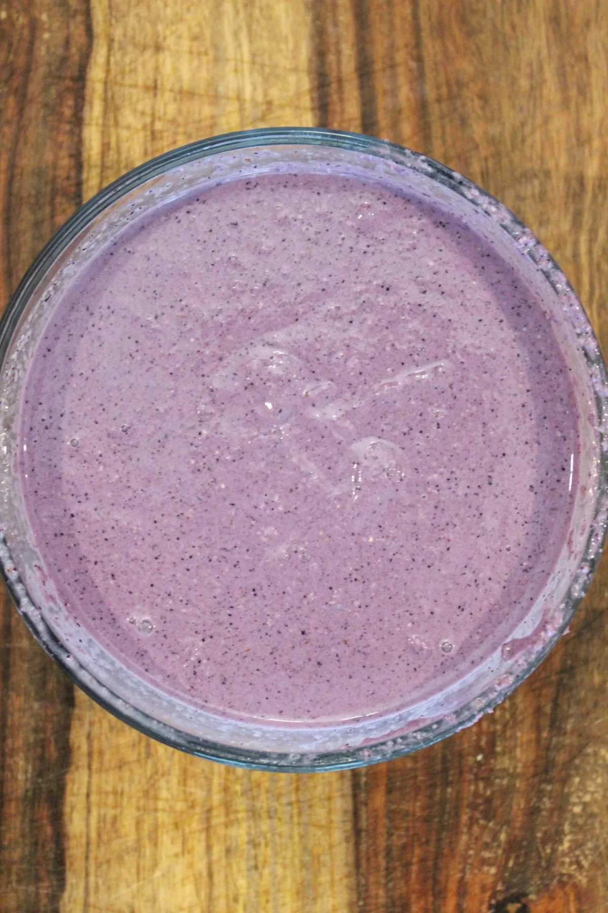 A glass bowl filled with smooth purple overnight oatmeal batter is placed on a wooden surface. The batter has a few visible air bubbles, suggesting it's freshly mixed.