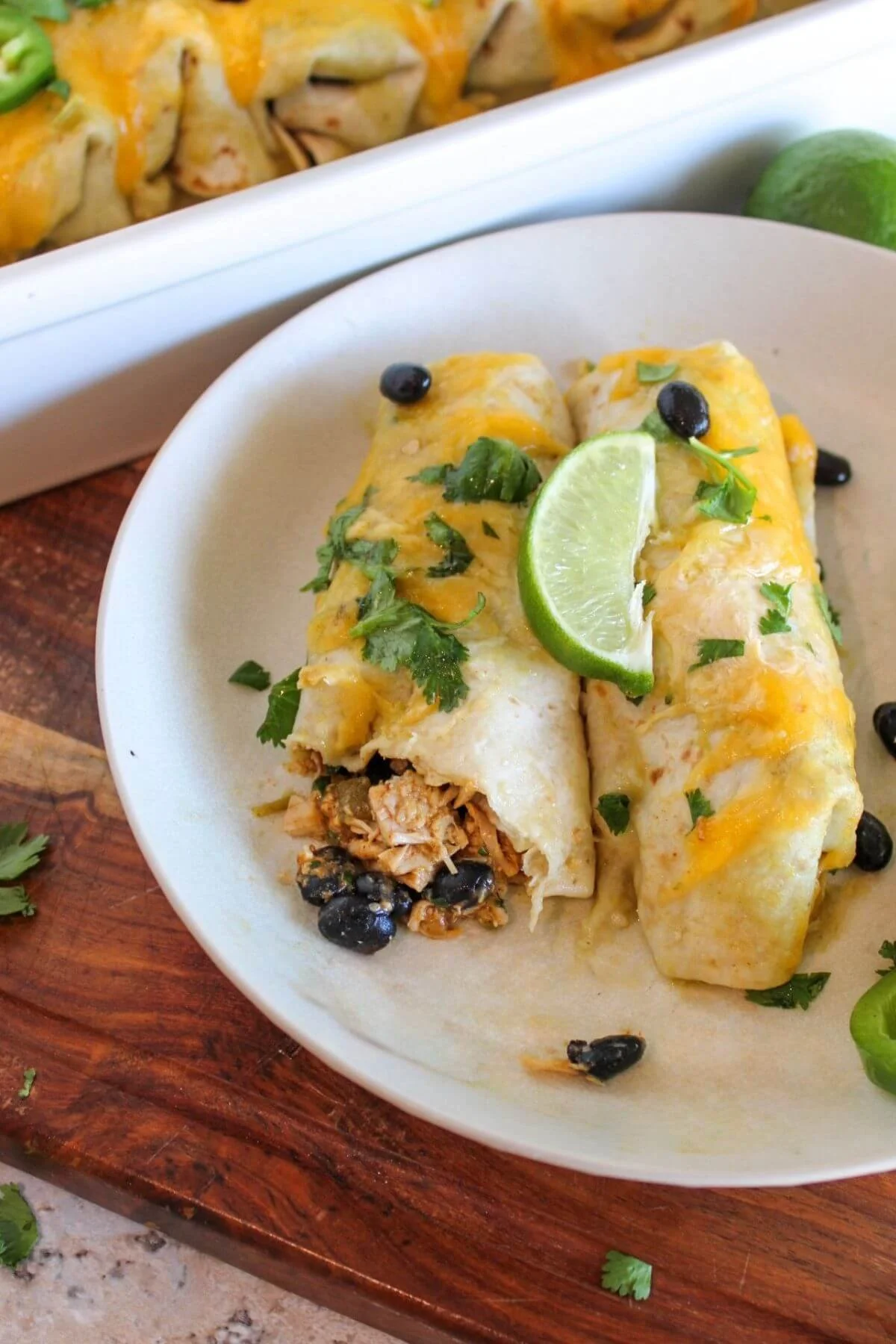 Two golden chicken-and-black-bean enchiladas topped with melted cheese, cilantro, lime, and black beans on a white plate.