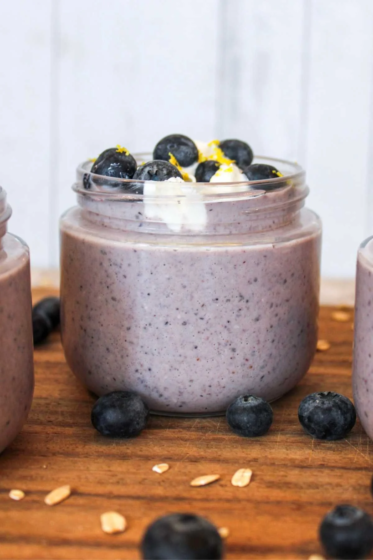 A creamy blueberry overnight oatmeal recipe in a glass jar topped with fresh blueberries and lemon zest sits on a wooden surface, surrounded by oats and more blueberries.