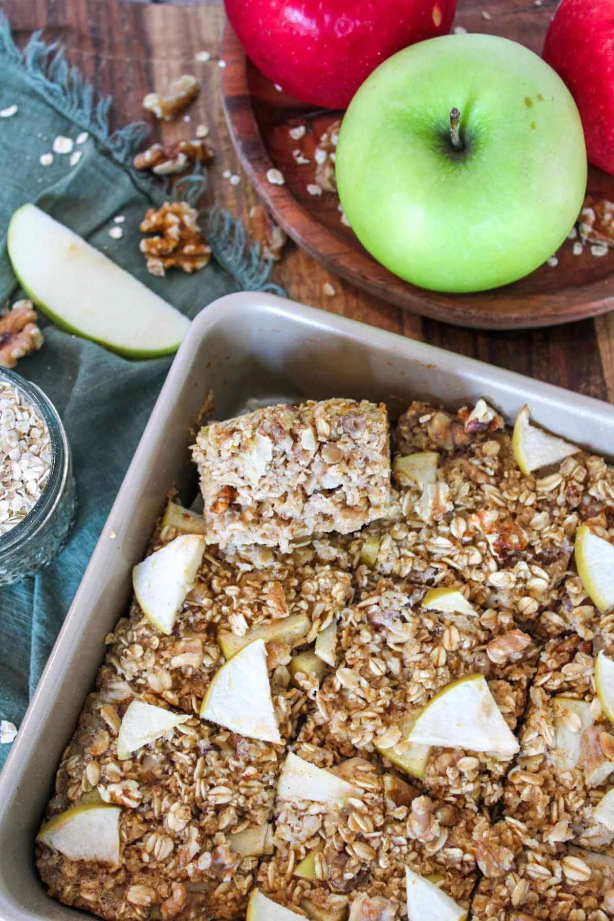 Overhead view of golden apple oat bars in a baking pan, with sliced green apple, red apples, walnuts, and oats on a rustic table.