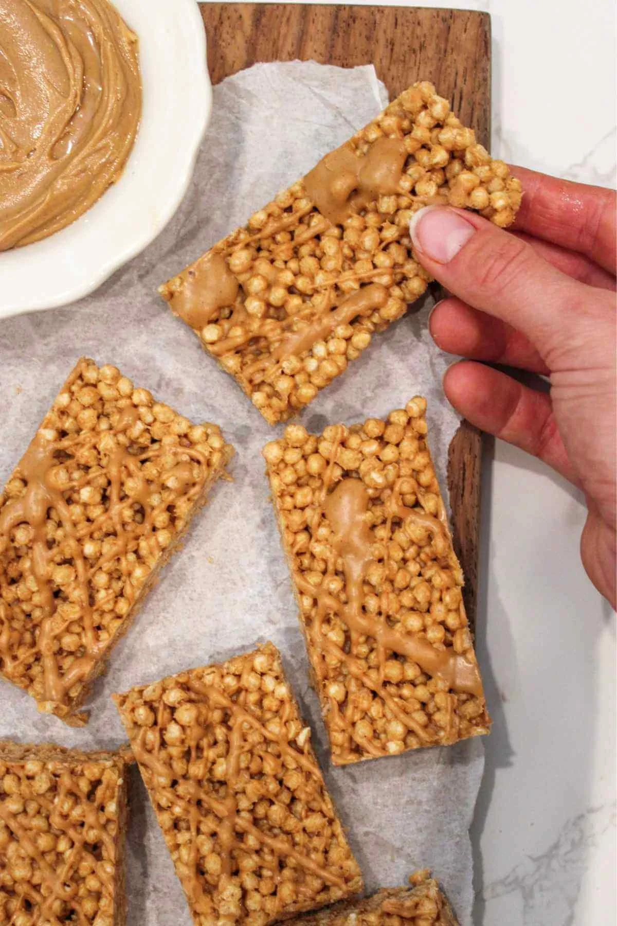 A hand holding a peanut butter granola bar over parchment paper and served on a wooden cutting board with a bowl of peanut butter next to the bars.