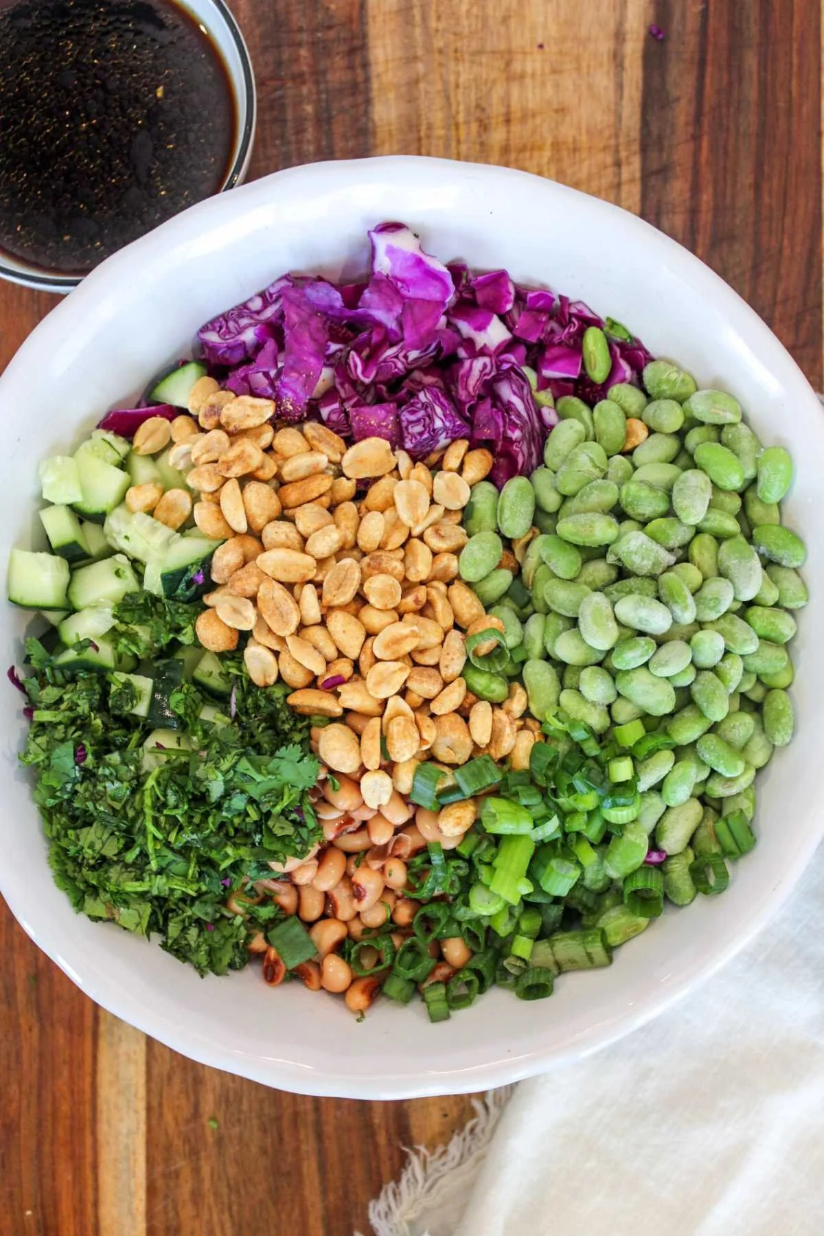 A vibrant salad in a white bowl with purple cabbage, edamame, peanuts, cucumber, cilantro, and green onions. A small dish of dark soy dressing is nearby.