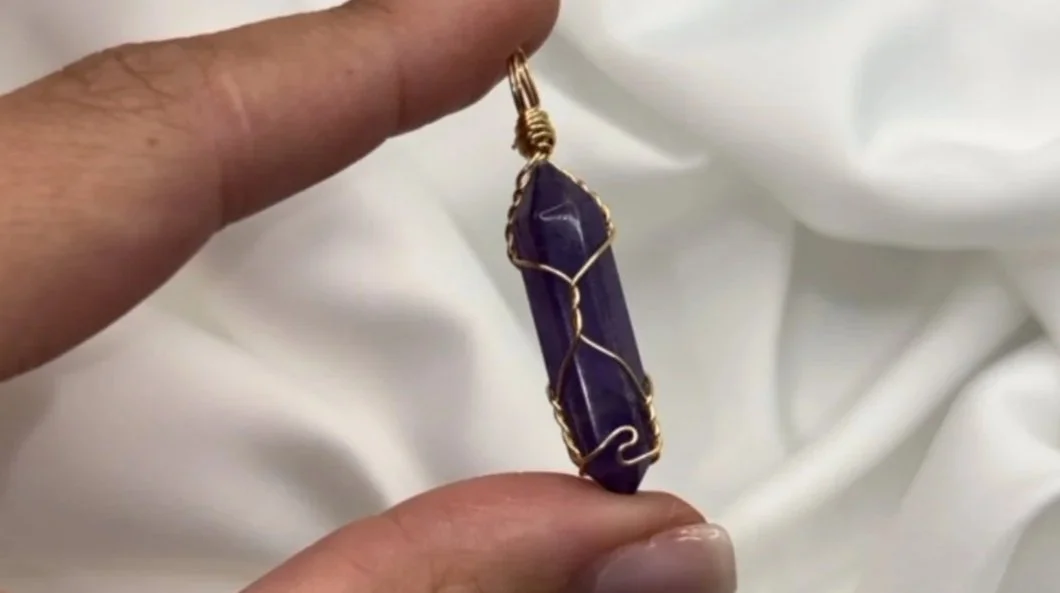 Purple crystal pendant with gold wire wrapping held against white fabric background.