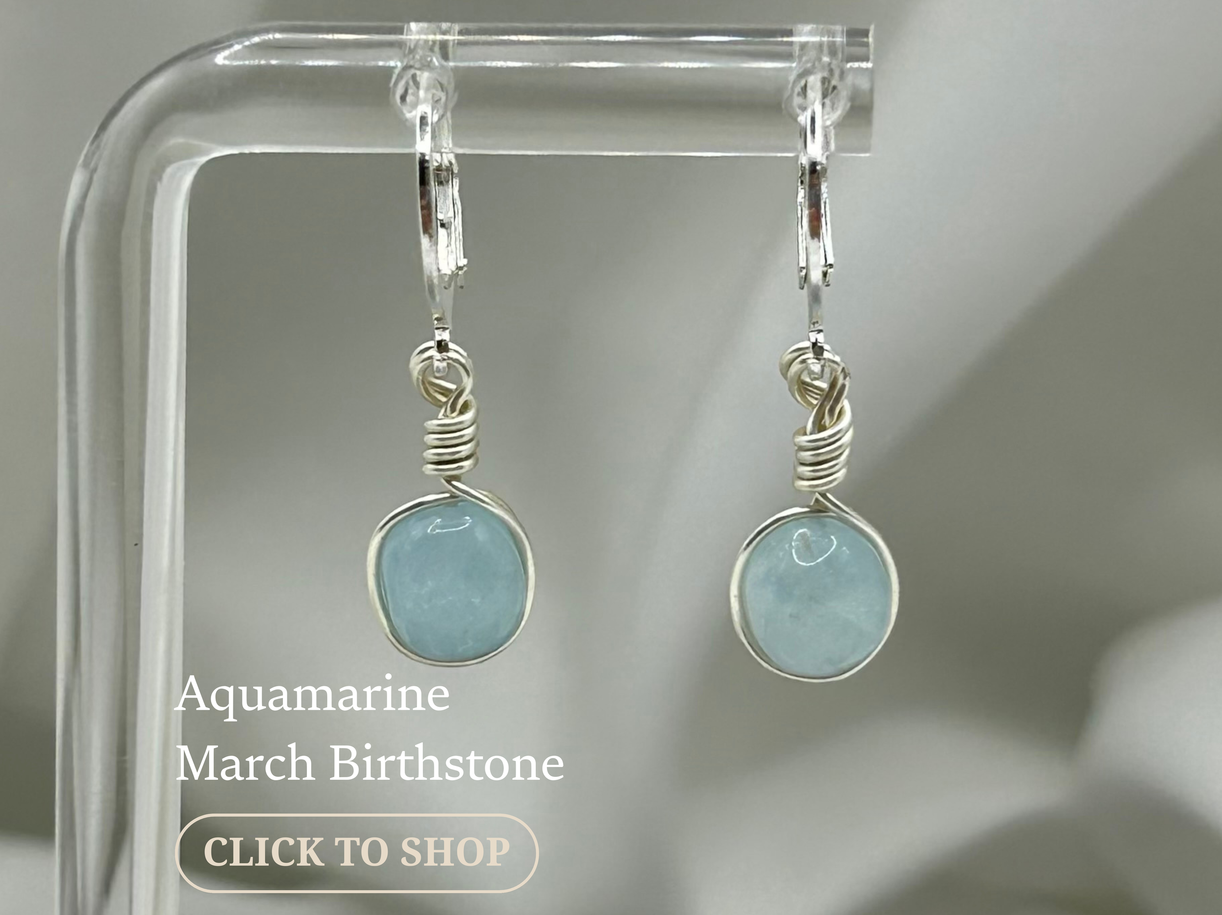 Pair of silver earrings with aquamarine stones hanging from hooks on a clear display stand, with text indicating they are March birthstone jewelry.