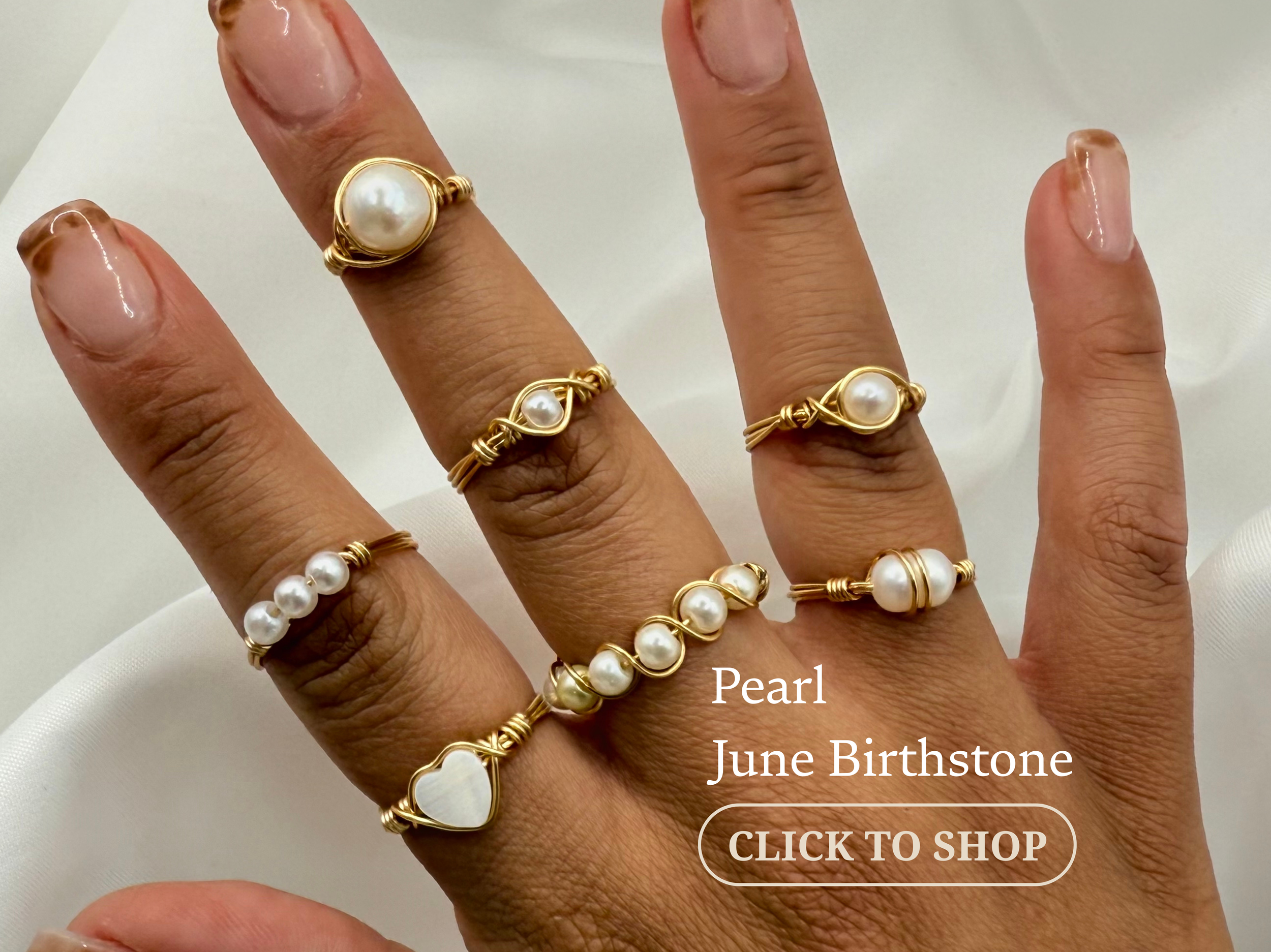 Multiple gold rings with pearl and shell accents on fingers, on a hand with light brown skin and manicured nails, with white fabric background.