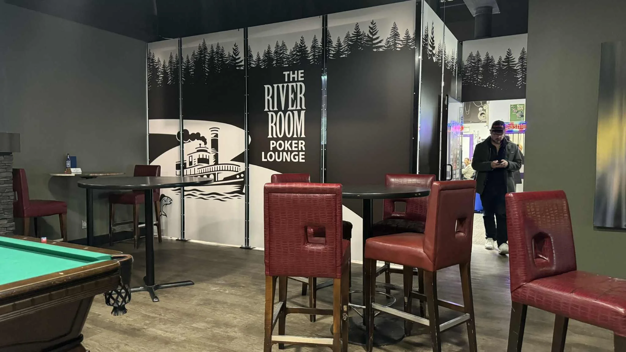 Interior of a lounge area with red chairs, black tables, and a large sign that reads 'The River Room Poker Lounge' featuring a lake and forest graphic. Trip Aces Poker Tour The River Room Poker Lounge at Flea Whiskeys Winnipeg Manitoba