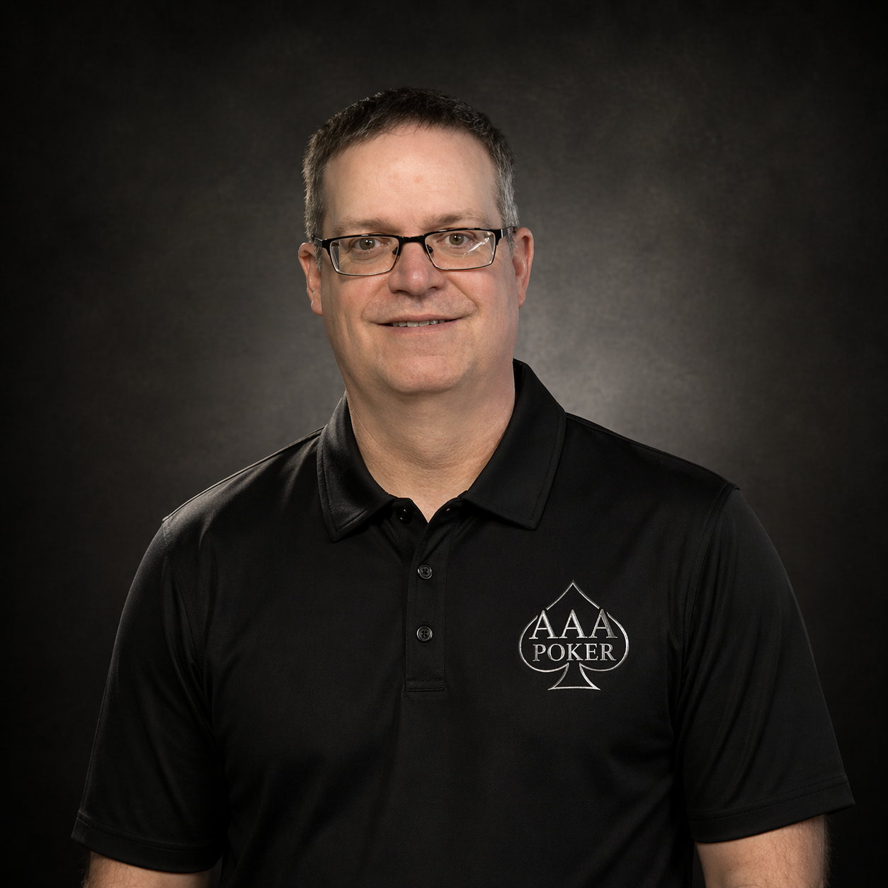 A man with short dark hair, glasses, and a black polo shirt with an AAA Poker logo, smiling against a dark background.