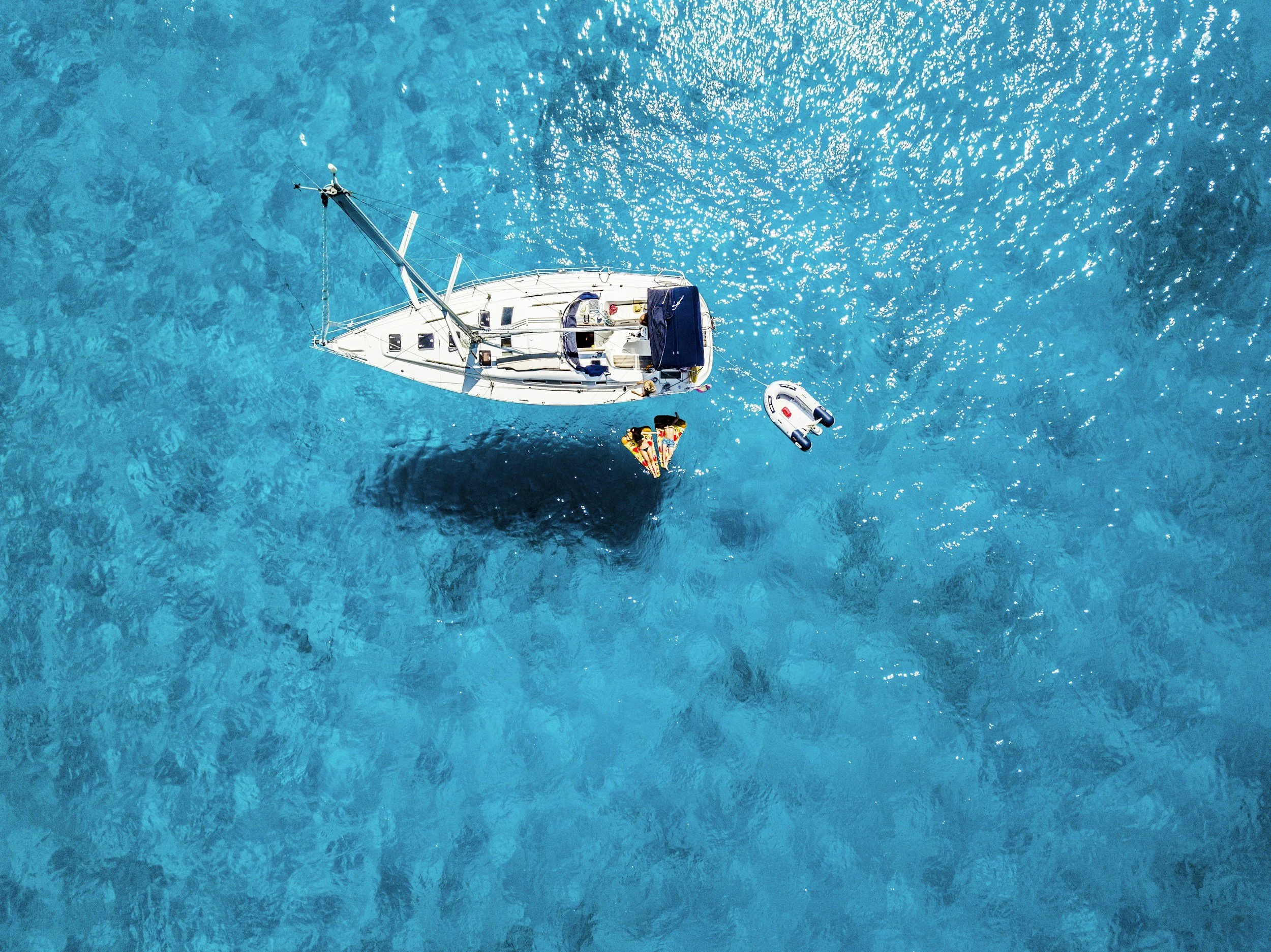 Aerial view of a white sailboat with a dark blue cover, floating on clear blue water, with a person sunbathing on a yellow and red float nearby.