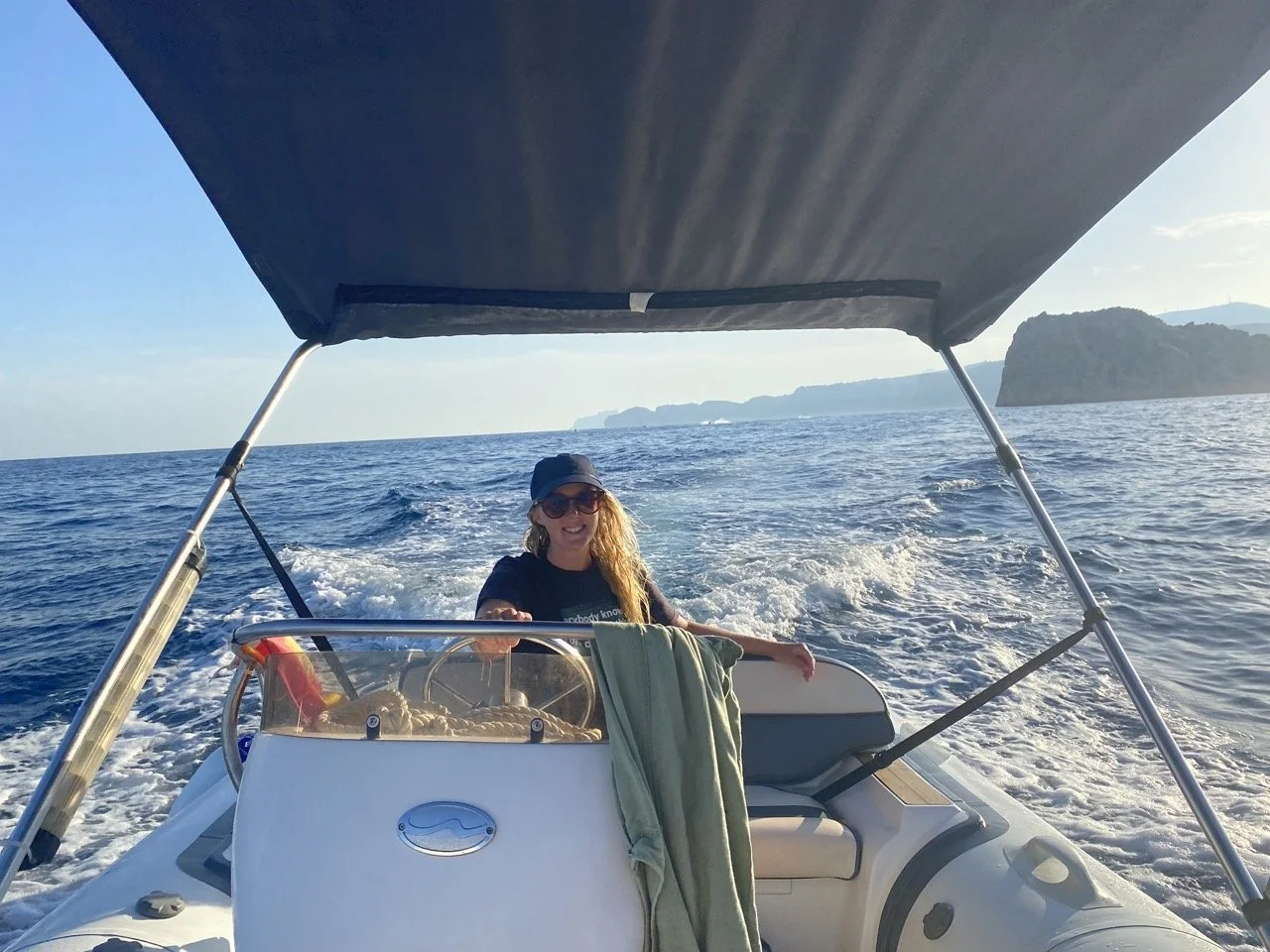 A woman on a small boat smiling, wearing sunglasses and a cap, with water and distant landforms in the background.