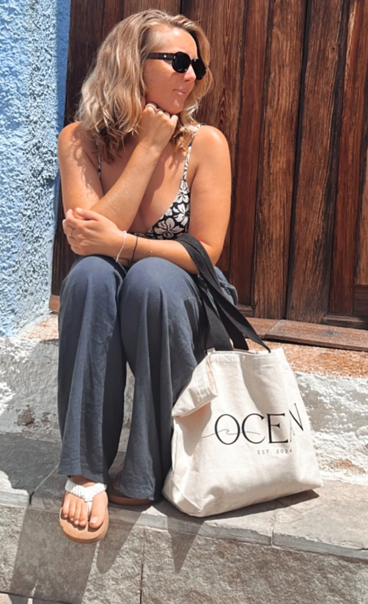 A woman with wavy blonde hair wearing black sunglasses, a patterned tank top, and loose pants, sitting on a stone bench near a wooden wall and blue wall, holding a bag with 'OCEAN' written on it.