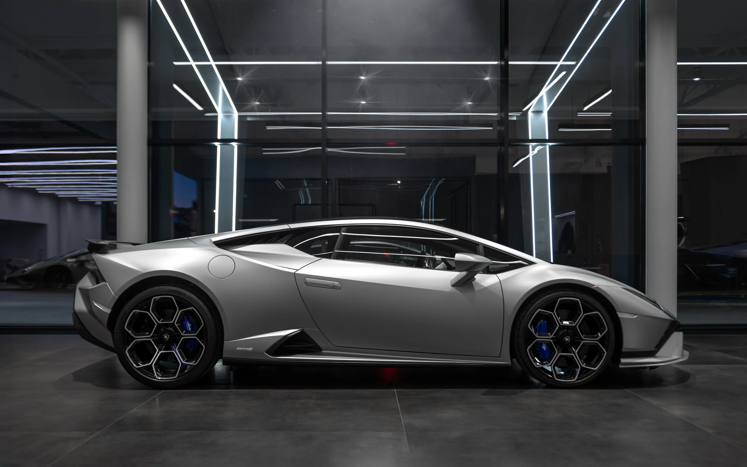 A silver Lamborghini sports car displayed indoors, with a sleek, modern design and black hexagonal wheel rims, in a showroom with reflective glass walls and modern lighting.