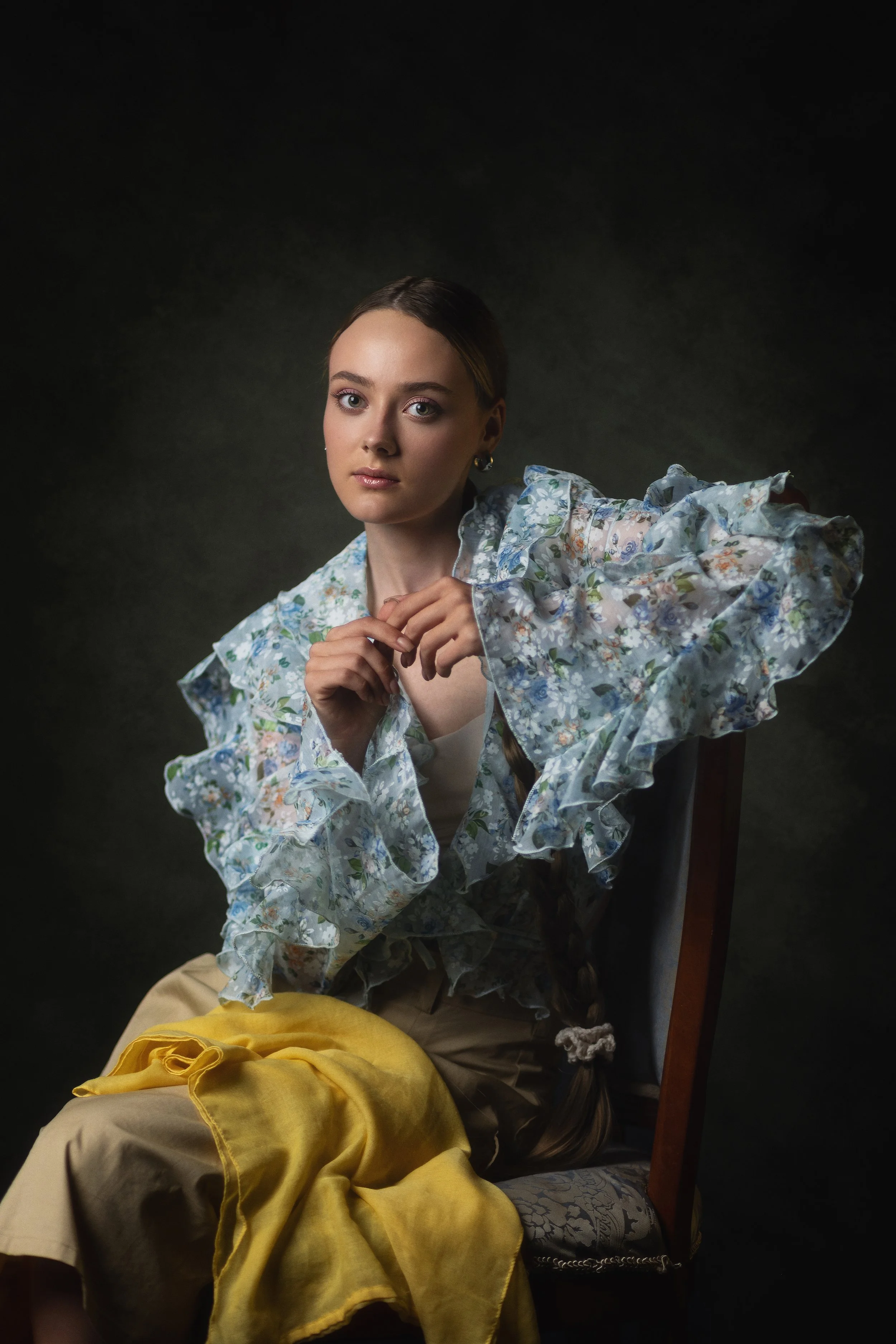 Young woman with fair skin and brown hair styled in a braid, wearing a floral ruffled blouse and yellow skirt, sitting on a wooden chair against a dark background.