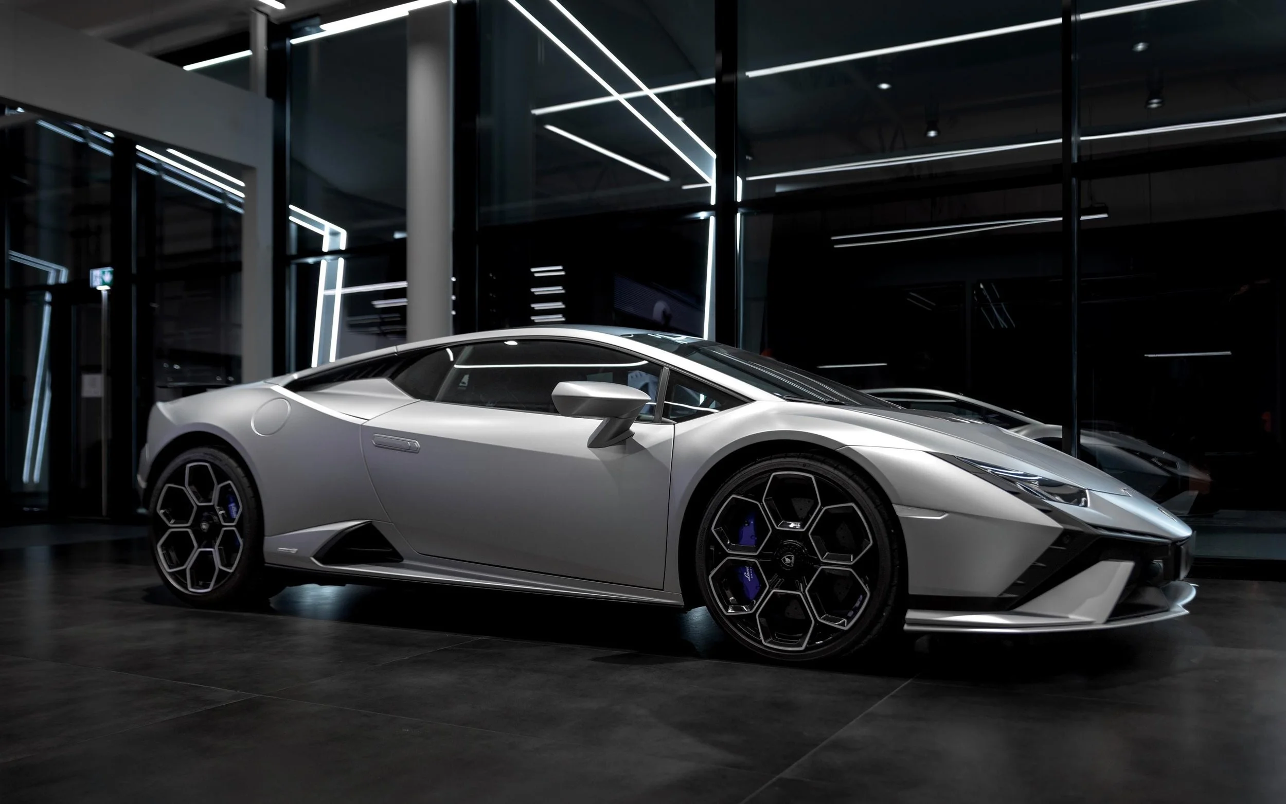 A silver Lamborghini sports car displayed in a modern, dark showroom with reflective glass walls and black floor tiles.