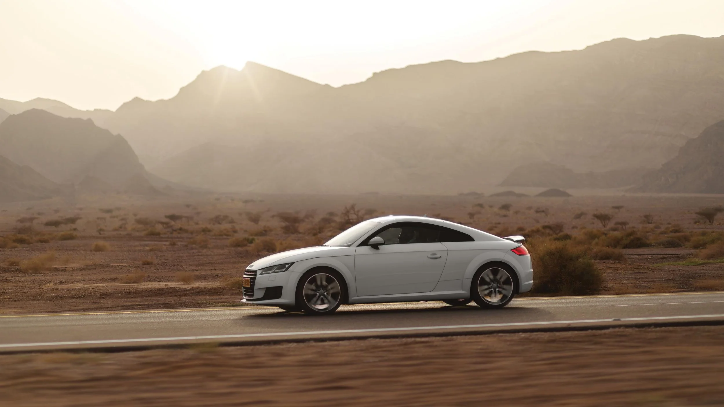 A silver sports car driving on a highway through a desert landscape with mountains in the background and the sun setting behind them.