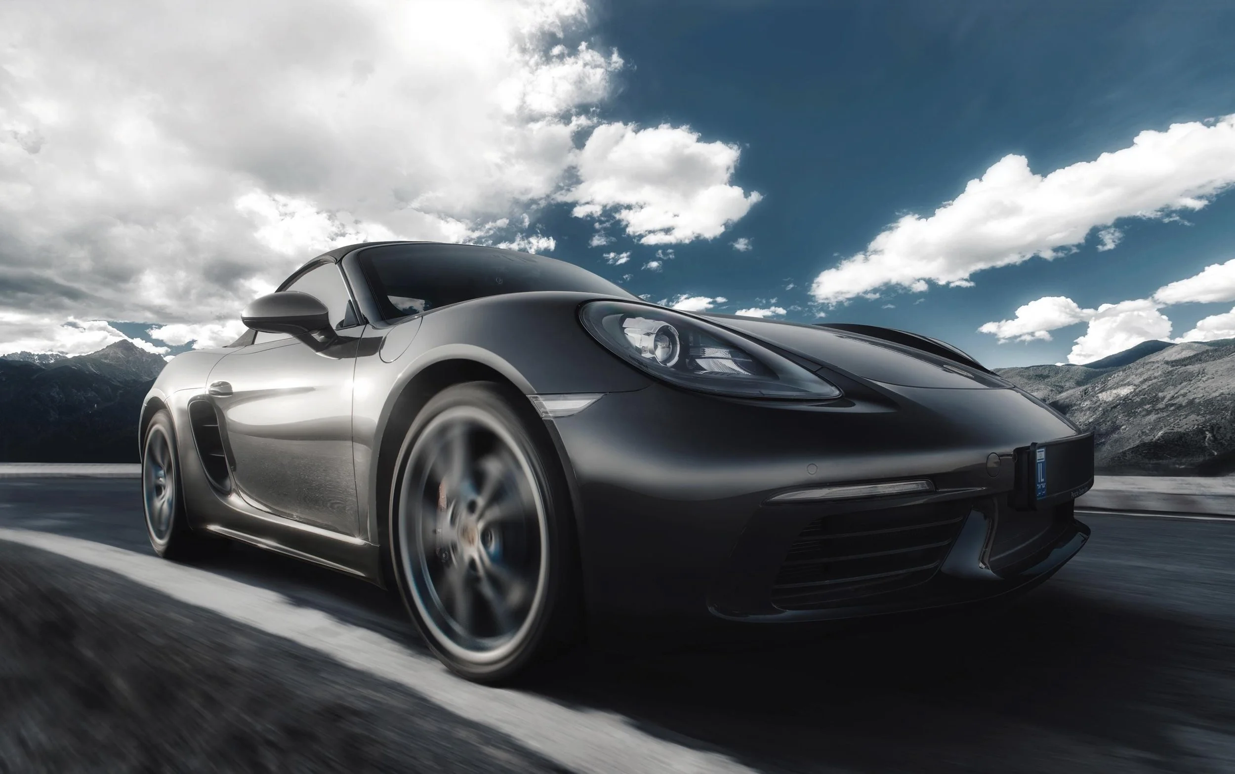 A sleek black sports car driving on a mountain road under a partly cloudy sky.