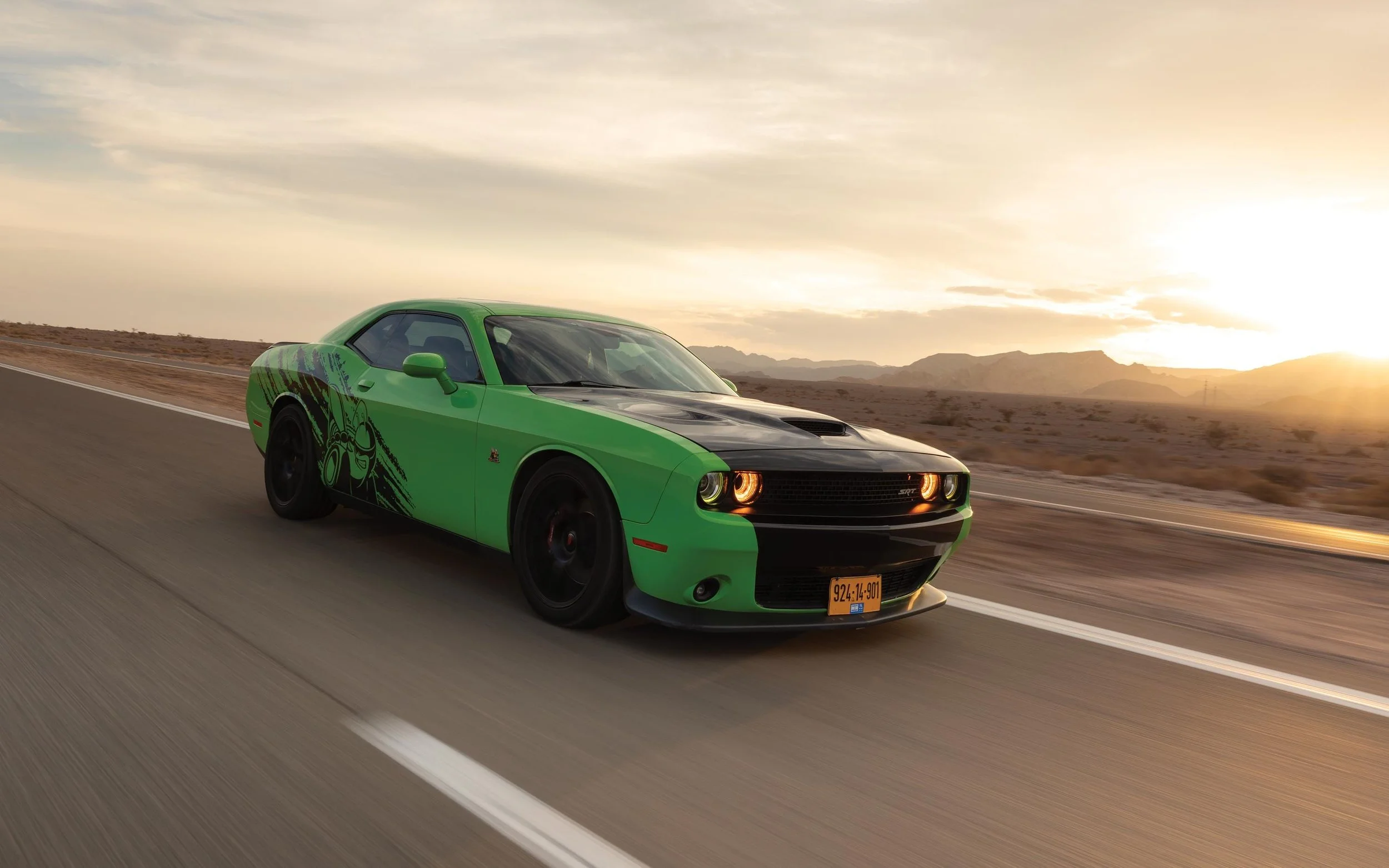 Green Dodge Challenger SRT driving on a desert highway at sunset.