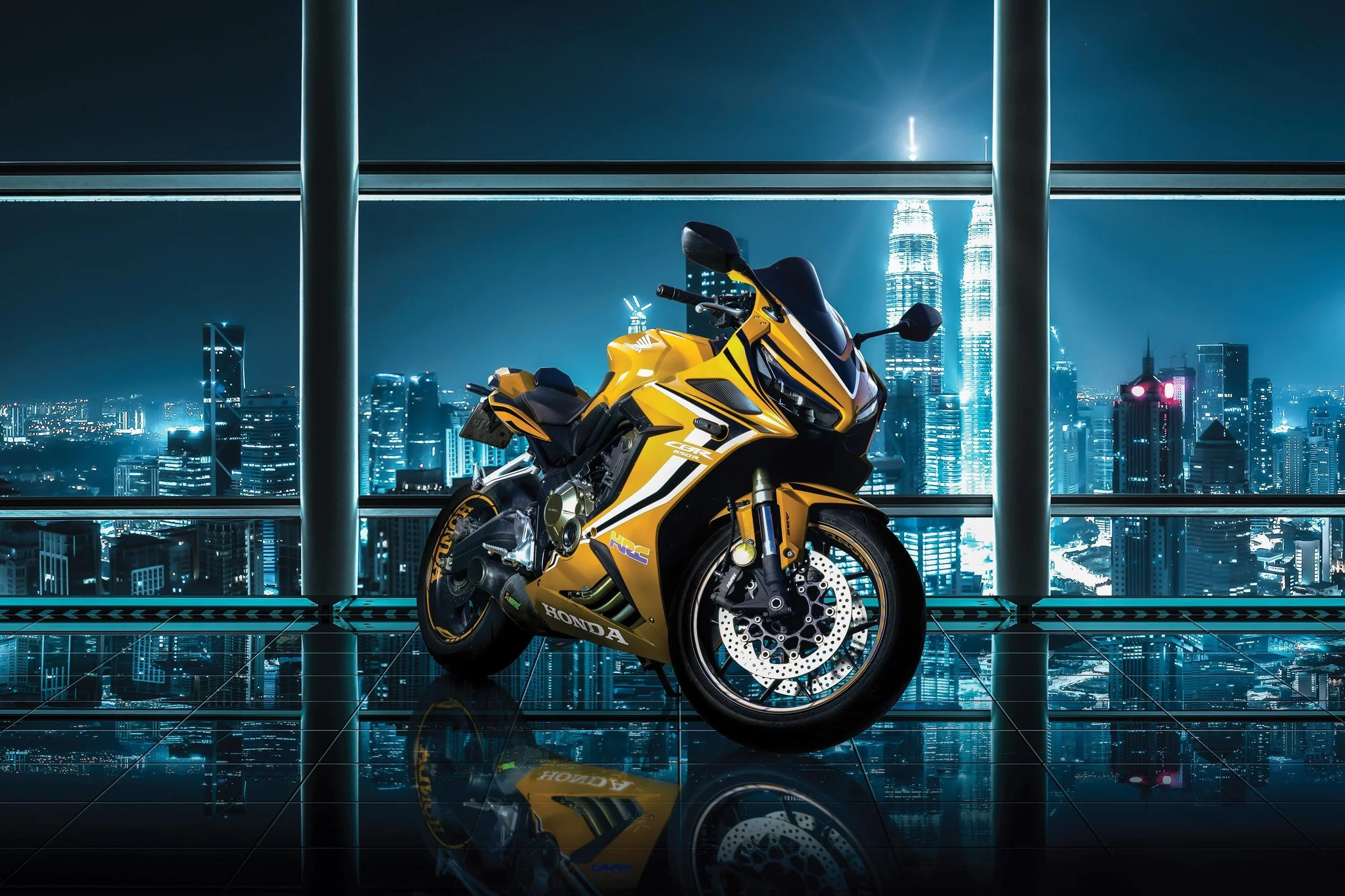 Yellow and black Honda sports motorcycle on a reflective tiled floor inside a high-rise building with large windows and a city skyline at night in the background.