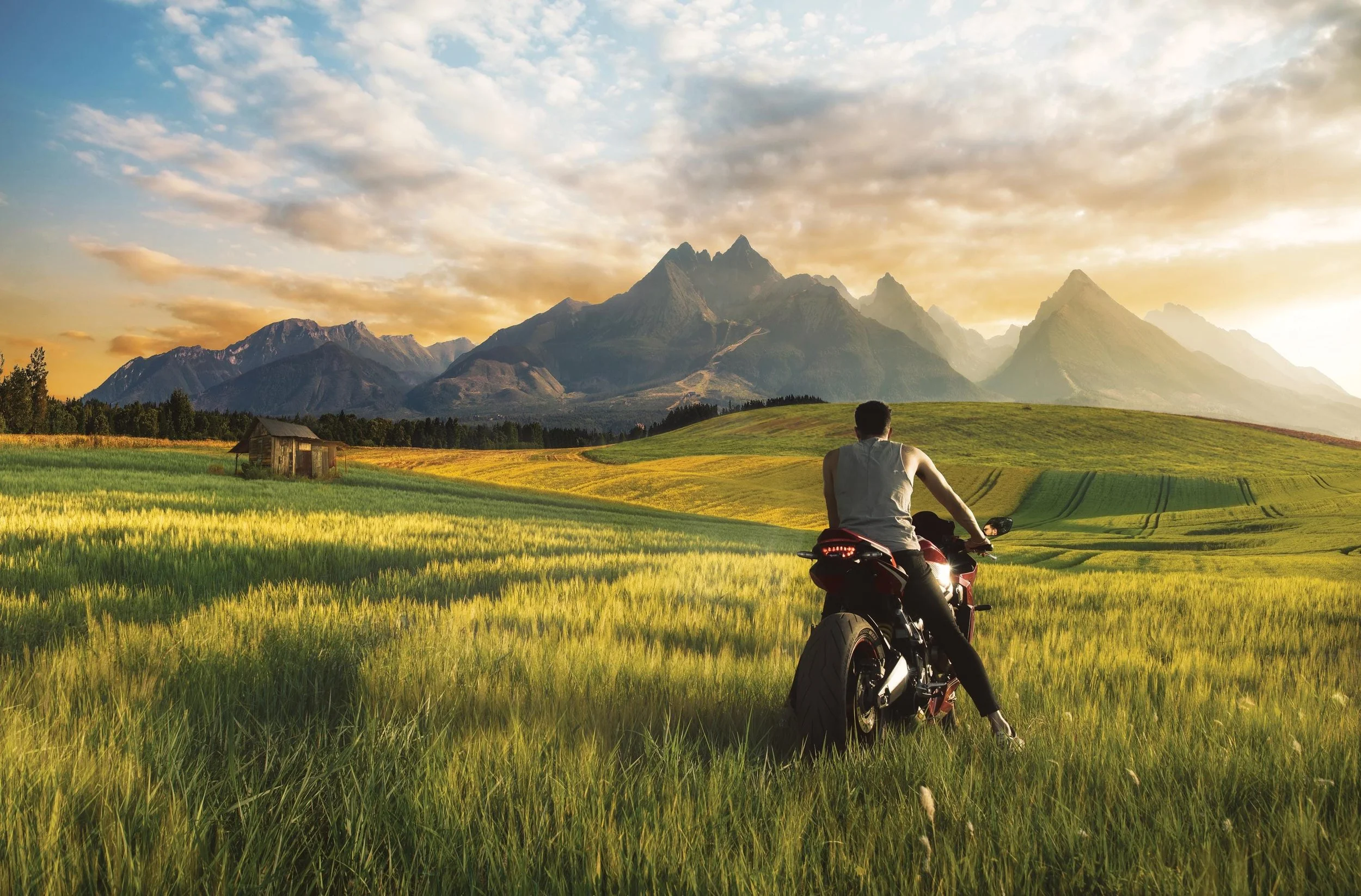 A man sitting on a motorcycle in a lush green field with mountains in the background and a sky with clouds at sunset.