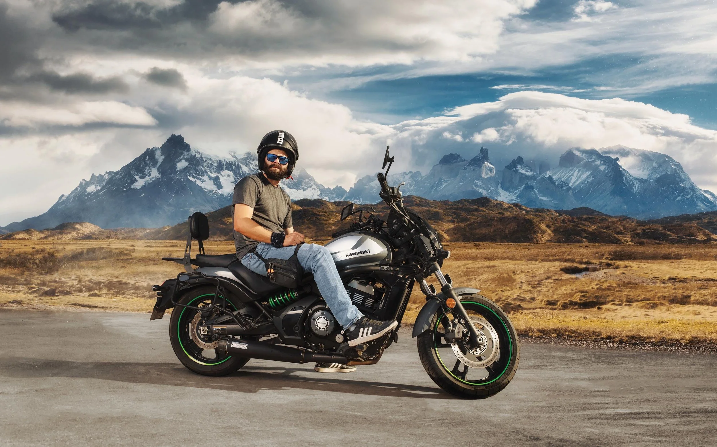 A man wearing a helmet and sunglasses sits on a black Kawasaki motorcycle on a deserted road with a mountainous landscape in the background.