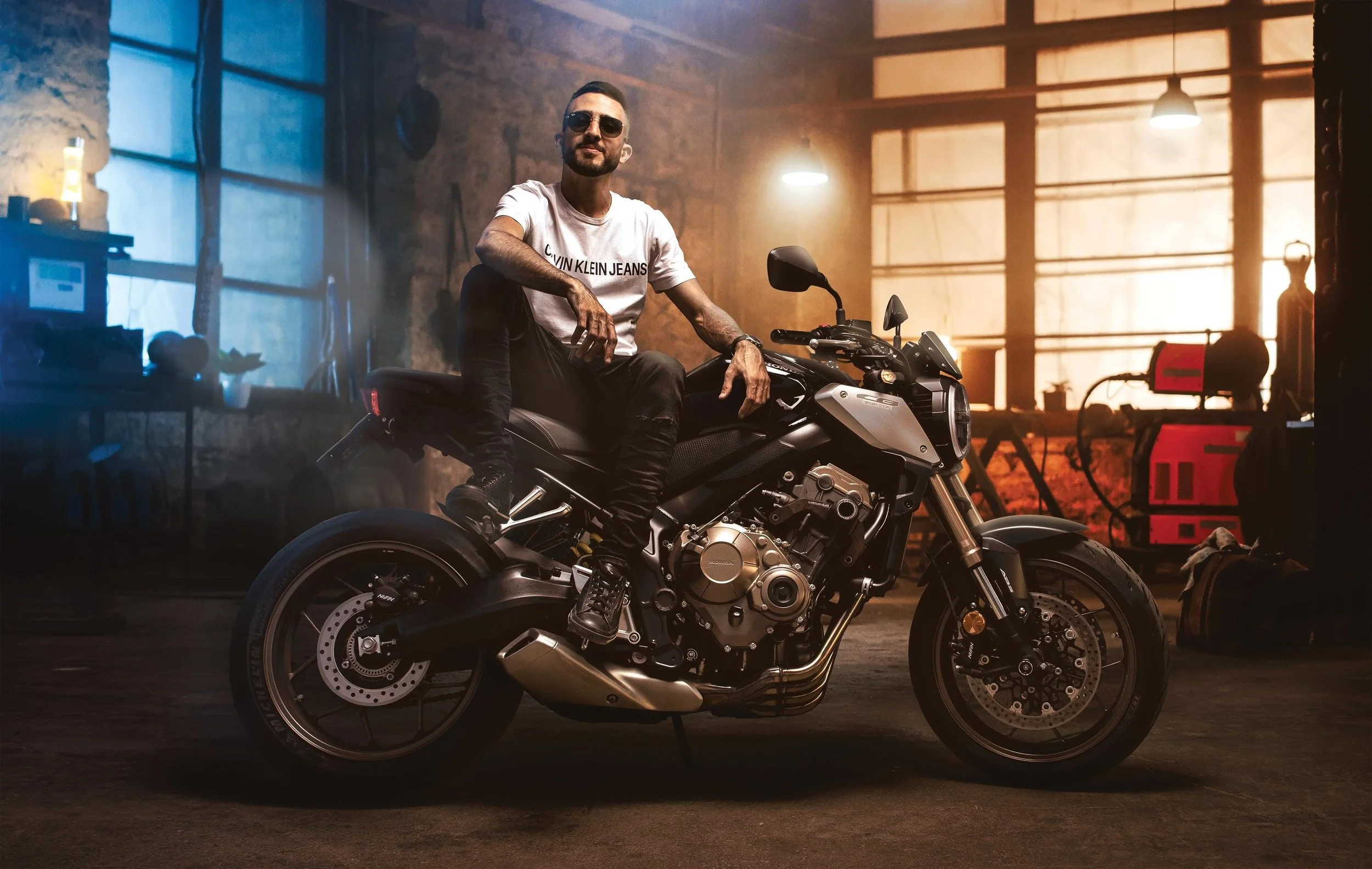 A man sitting on a black motorcycle inside a garage or workshop, with tools and equipment visible in the background and warm lighting.