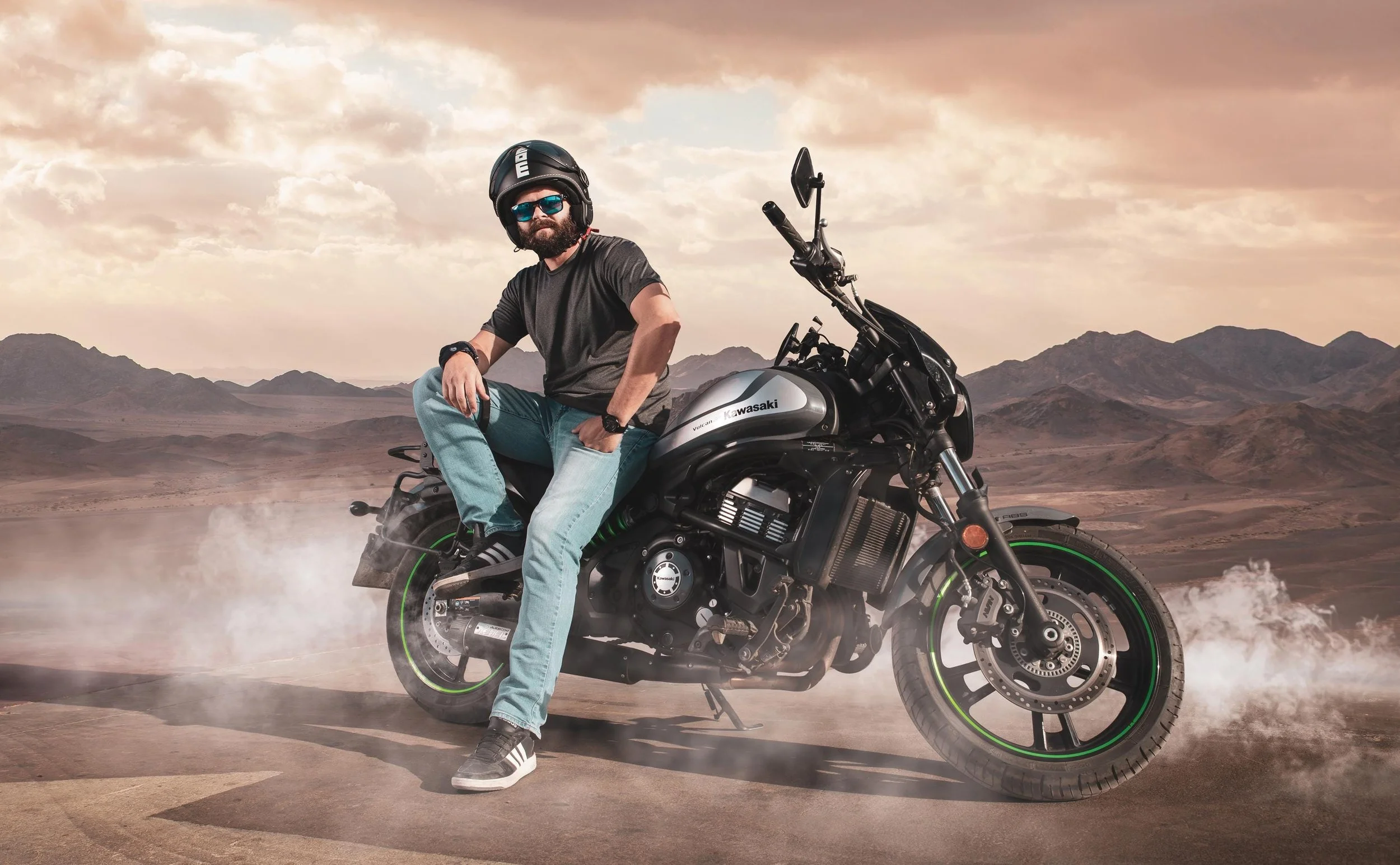 A man with a beard and sunglasses wearing a black helmet, black t-shirt, and blue jeans sitting on a black Kawasaki motorcycle in a desert landscape with mountains and cloudy sky.