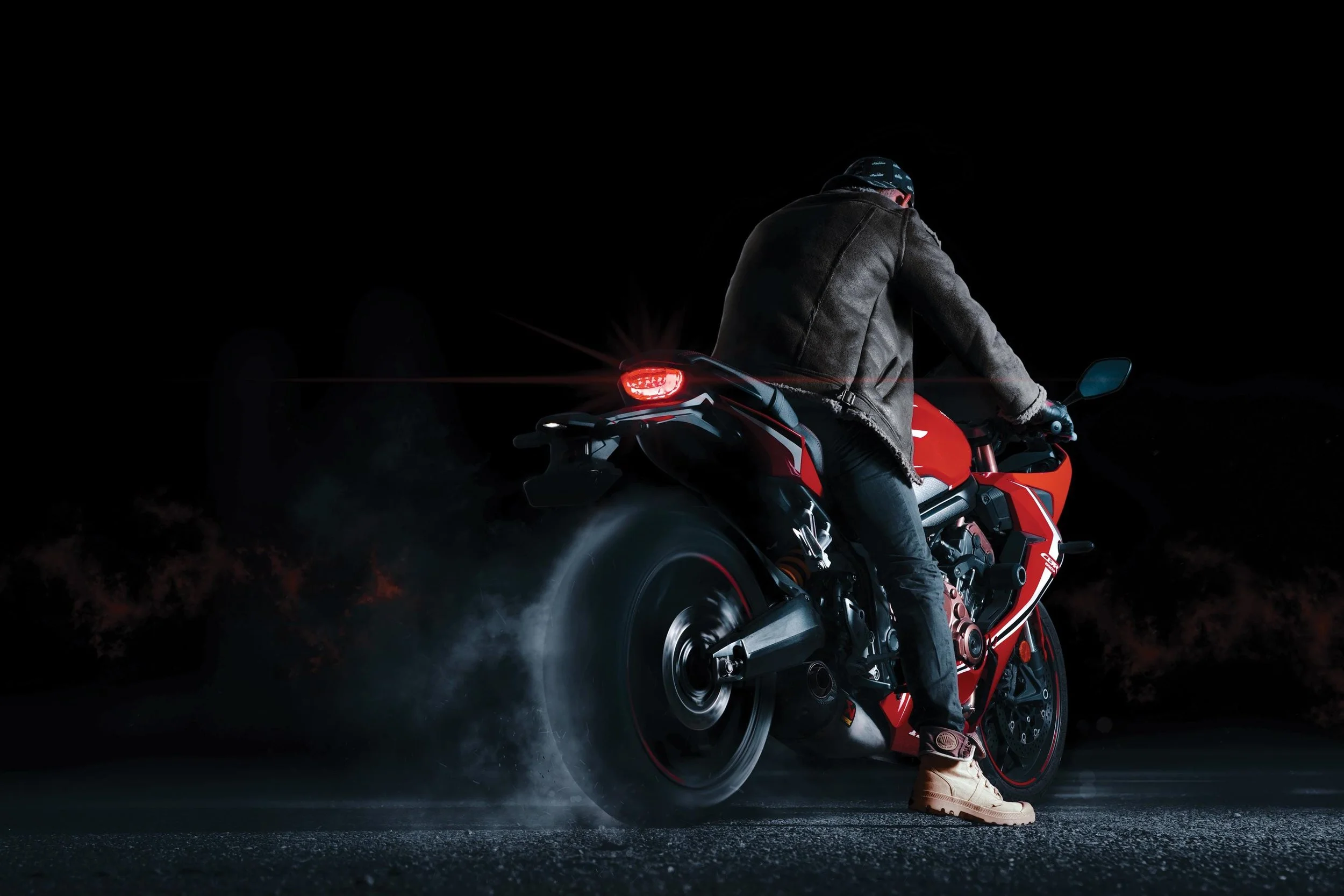 Person wearing a leather jacket and tan boots riding a red sport motorcycle on a dark road at night with smoke and dust around the rear tire.