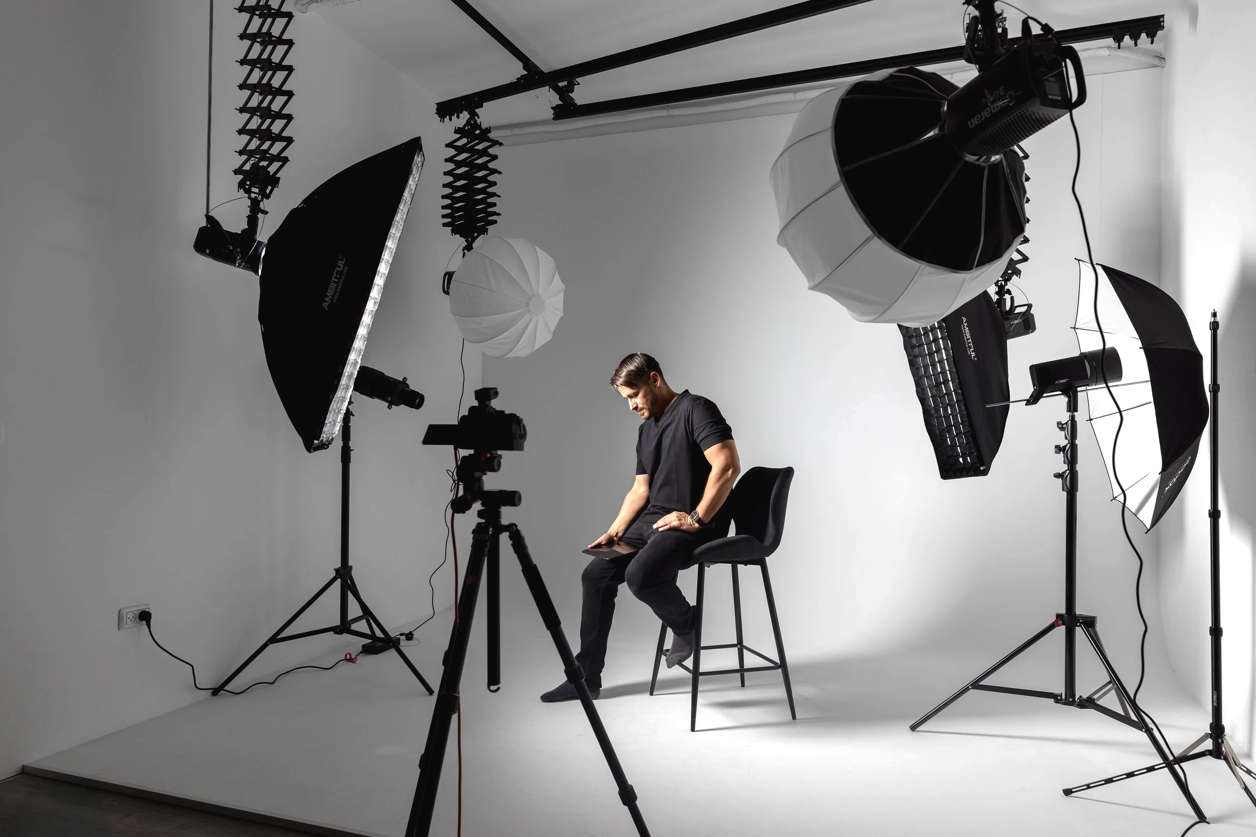 A man sitting on a chair in a photography studio, surrounded by professional lighting equipment including softboxes and umbrella lights, with a camera set up in front of him.