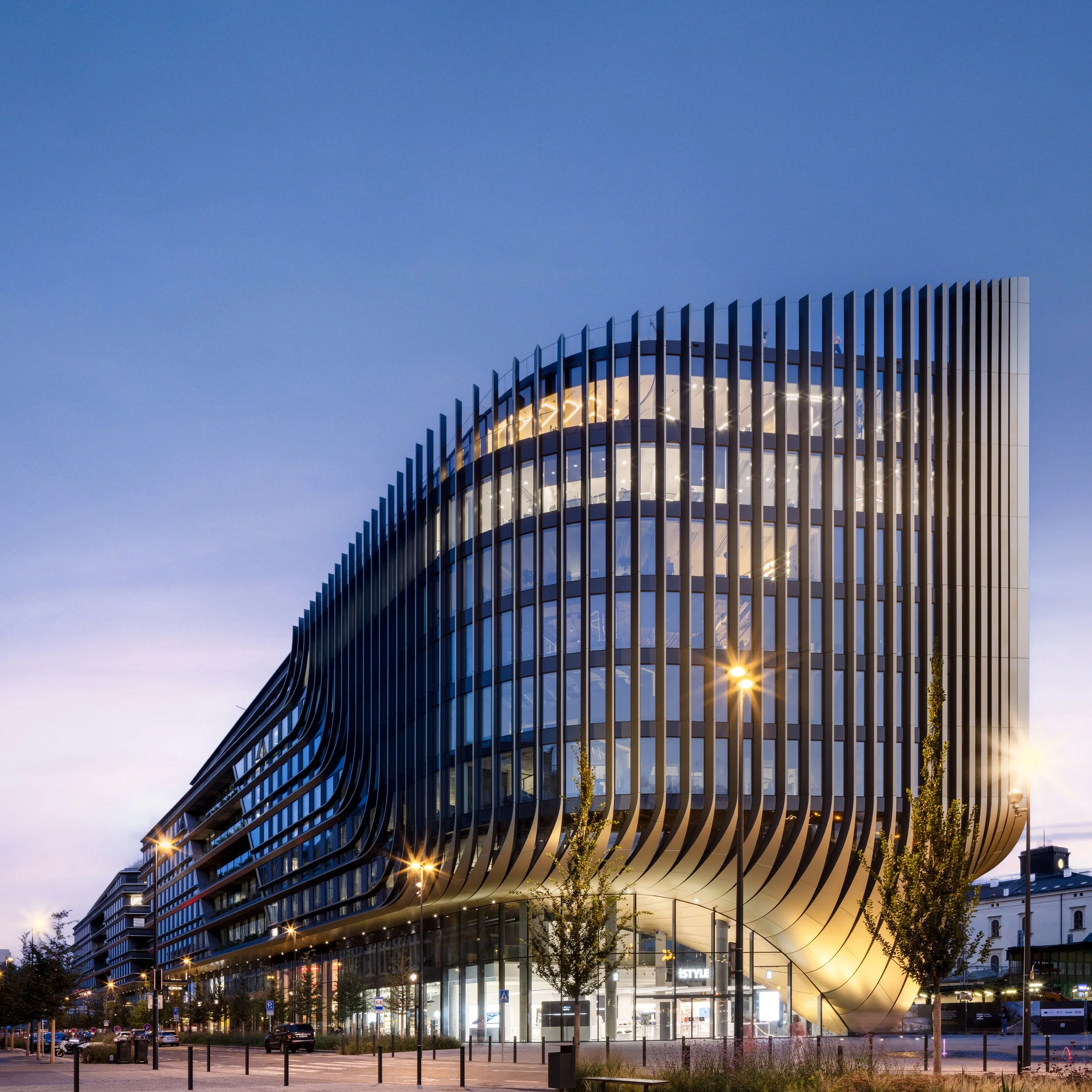 Modern multi-story building with curved glass facade and vertical black metal fins, illuminated at dusk, with streetlights, trees, and parked cars in front.