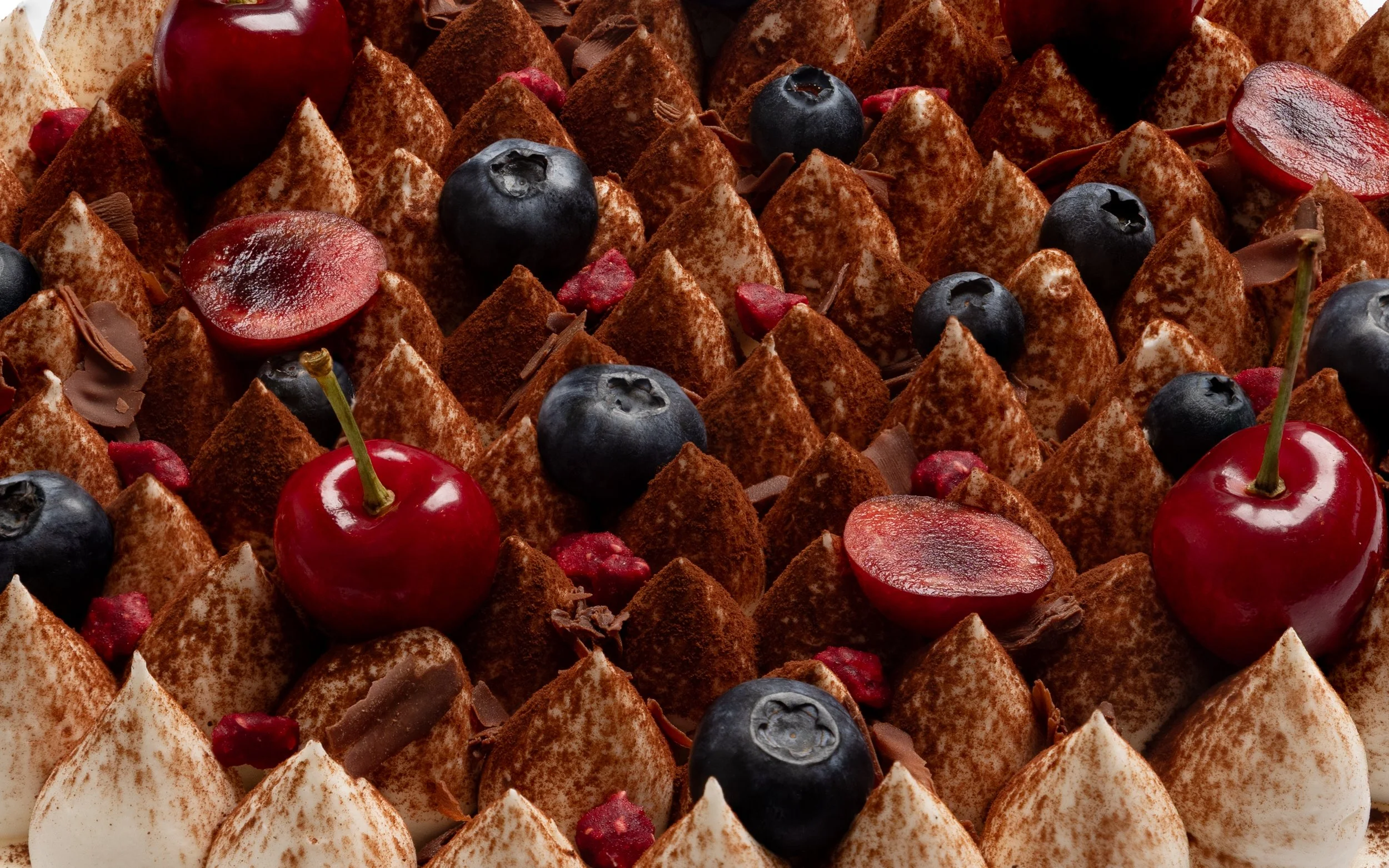 A close-up view of a cake decorated with cherries, blueberries, and chocolate shavings, with alternating white and chocolate frosting peaks dusted with cocoa powder.