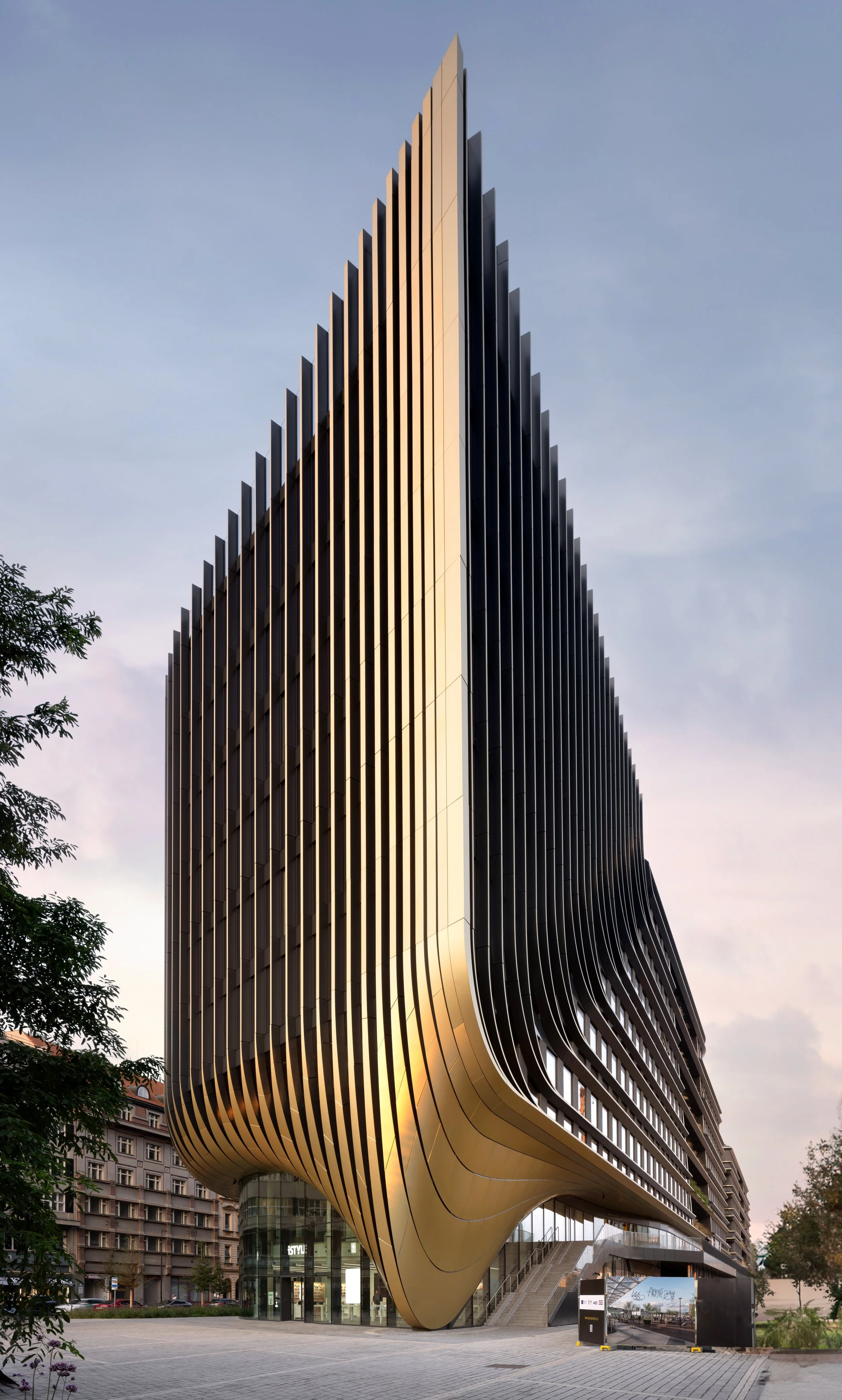 A modern high-rise building with a curved golden facade and vertical black slats, set against a dusk sky.