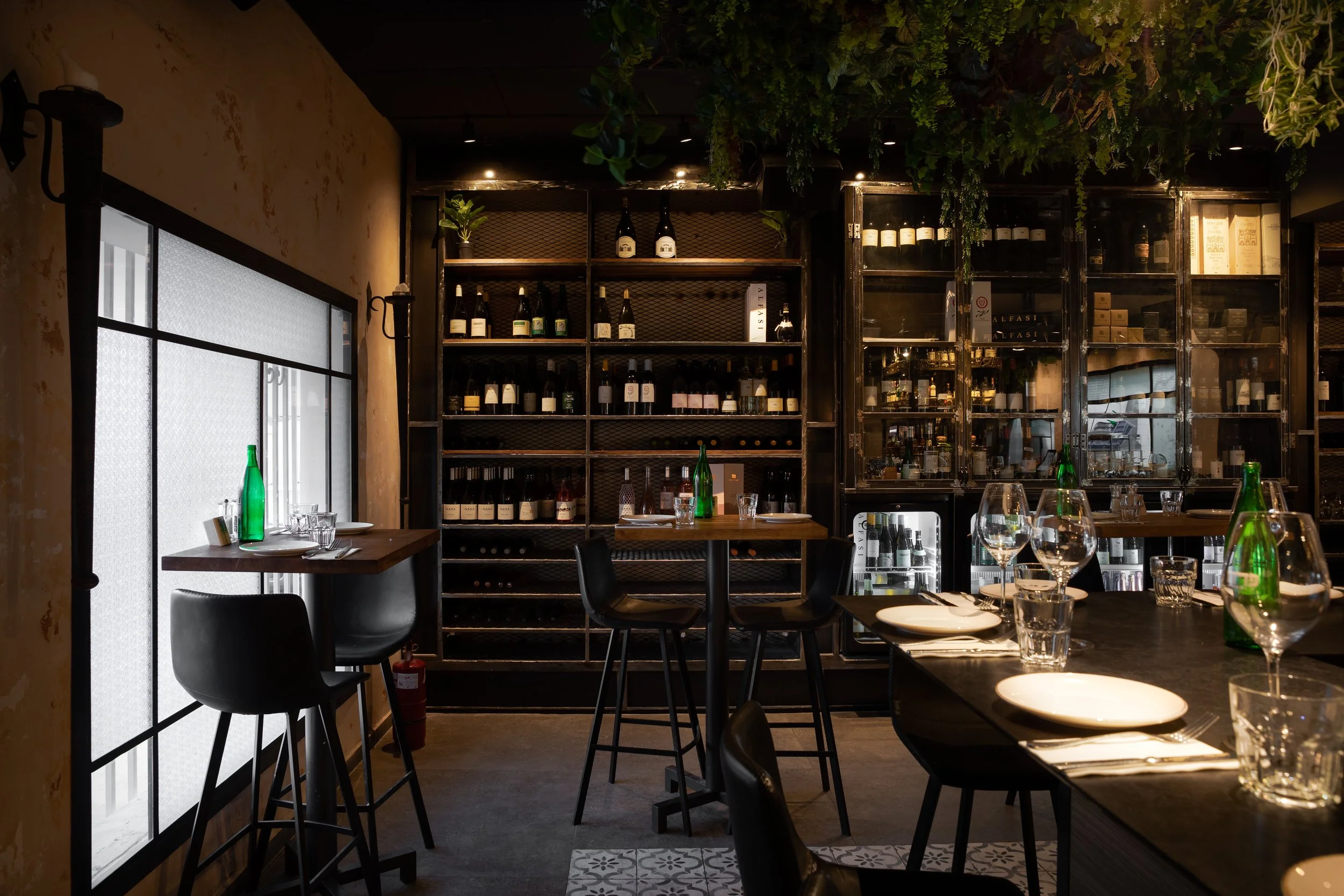 Interior of a cozy modern restaurant with black furniture, wine bottles on shelves, and set dining tables with plates, glasses, and green water bottles