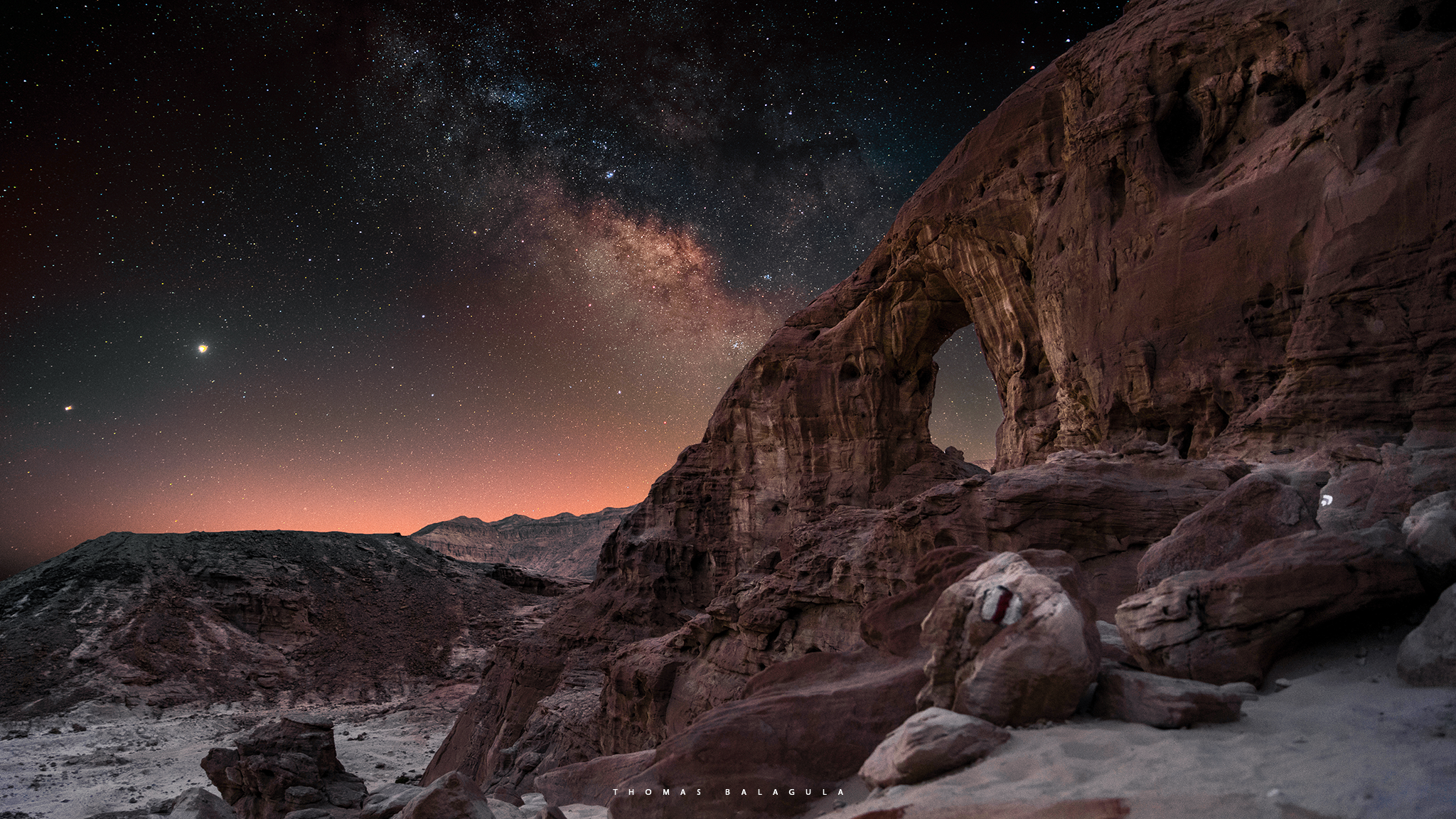 A desert scene at night with a natural rock arch formation, rocky terrain, and a starry sky featuring the Milky Way galaxy.