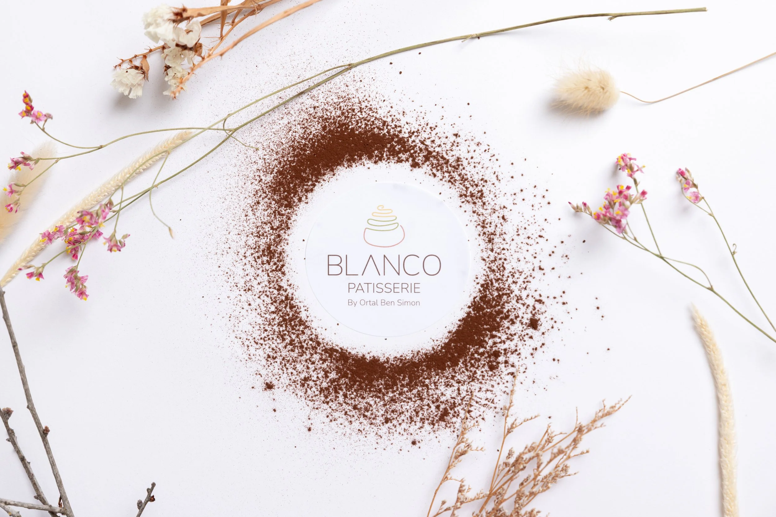A circular arrangement of cocoa powder around a white paper with 'Blanco Patisserie by Ortal Ben Simon' logo, surrounded by dried flowers on a white background.