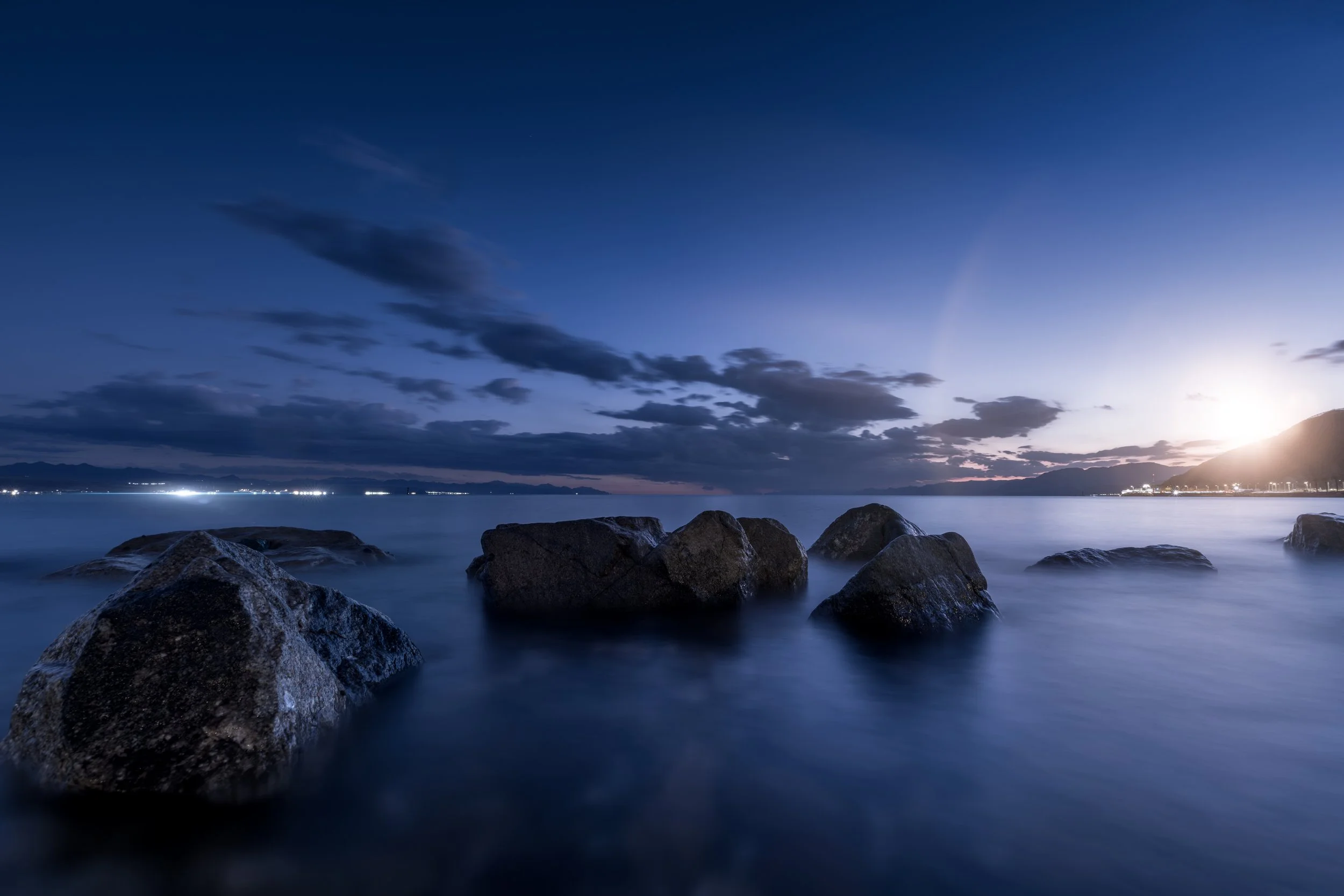 Nighttime seascape with large rocks in calm water, cloudy sky, and city lights in the distance, with the sun setting behind distant mountains.