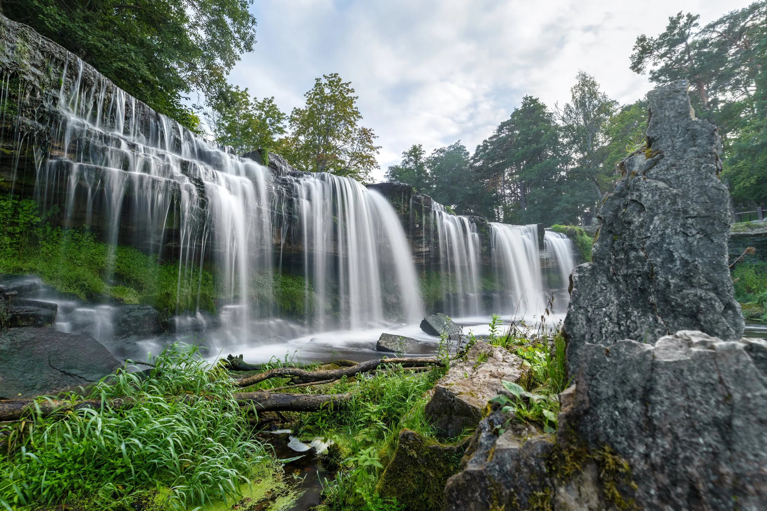 Waterfalls flowing over rocks amidst green trees and vegetation, with a partially cloudy sky above.