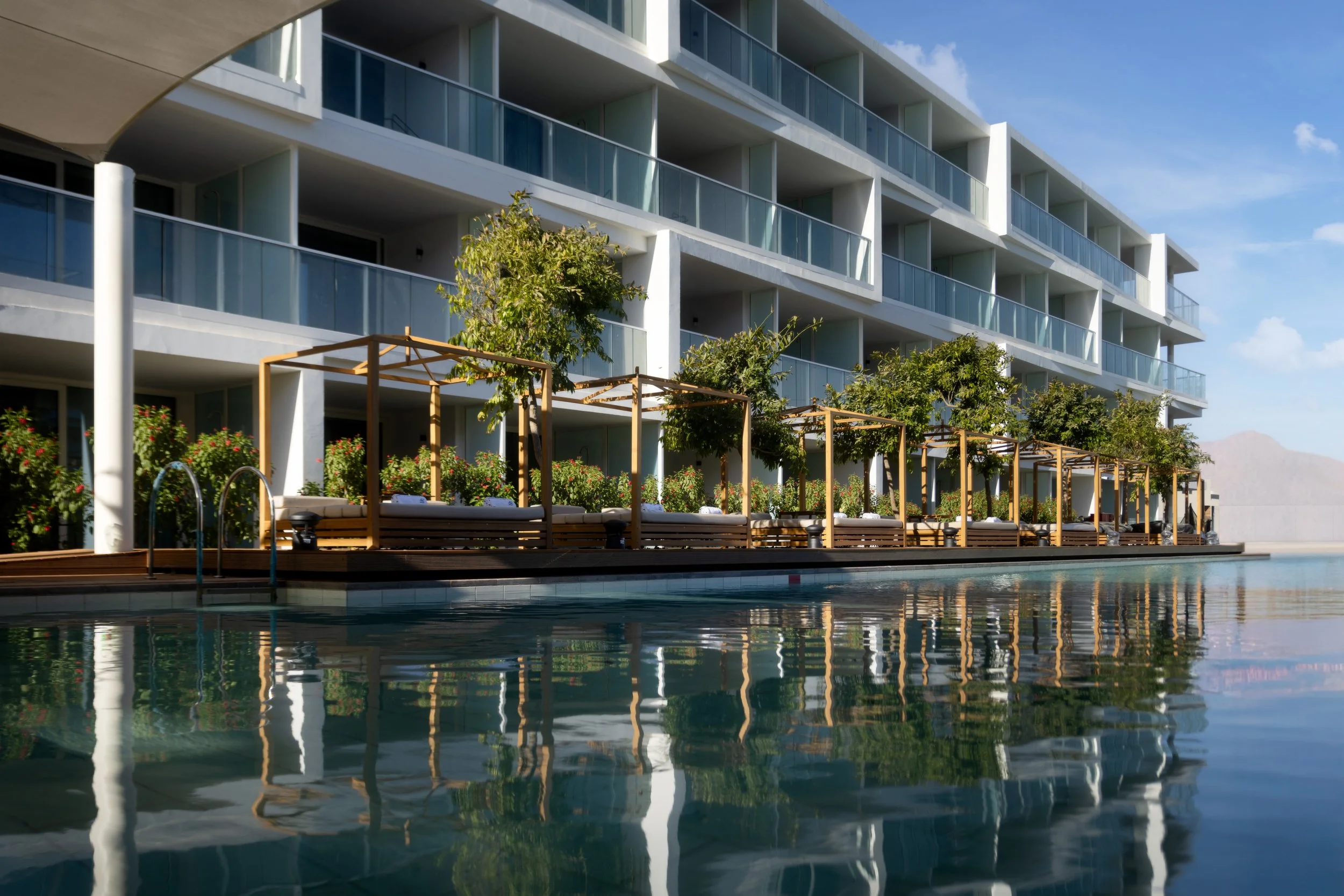 Modern multi-story hotel with balcony rooms, resting on a dockside with a swimming pool, lounge seating areas covered with wooden pergolas, and green trees under a blue sky.