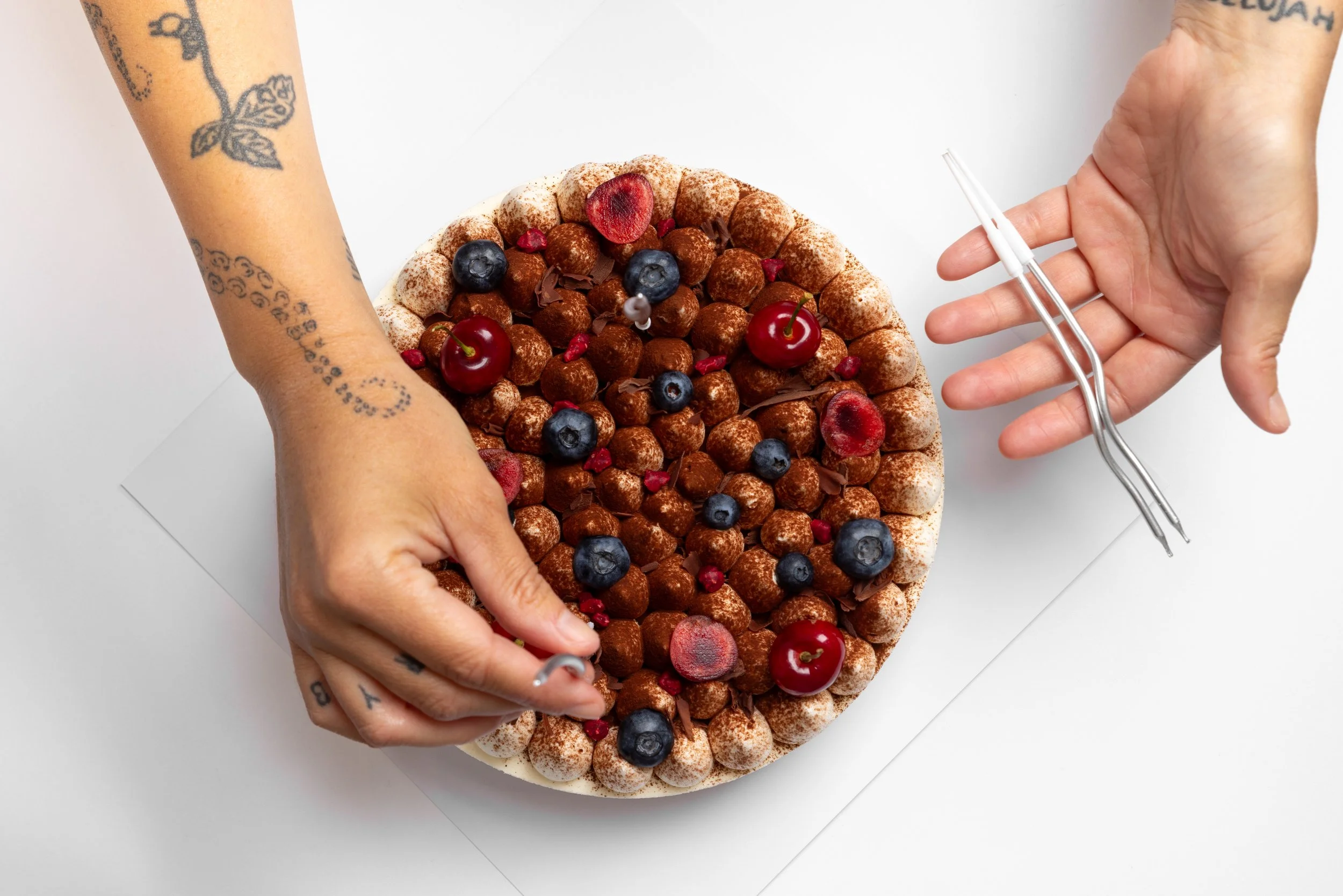 A person with tattoos is decorating a round cake with chocolate and berries using a fork and a cake tool, with a white background.