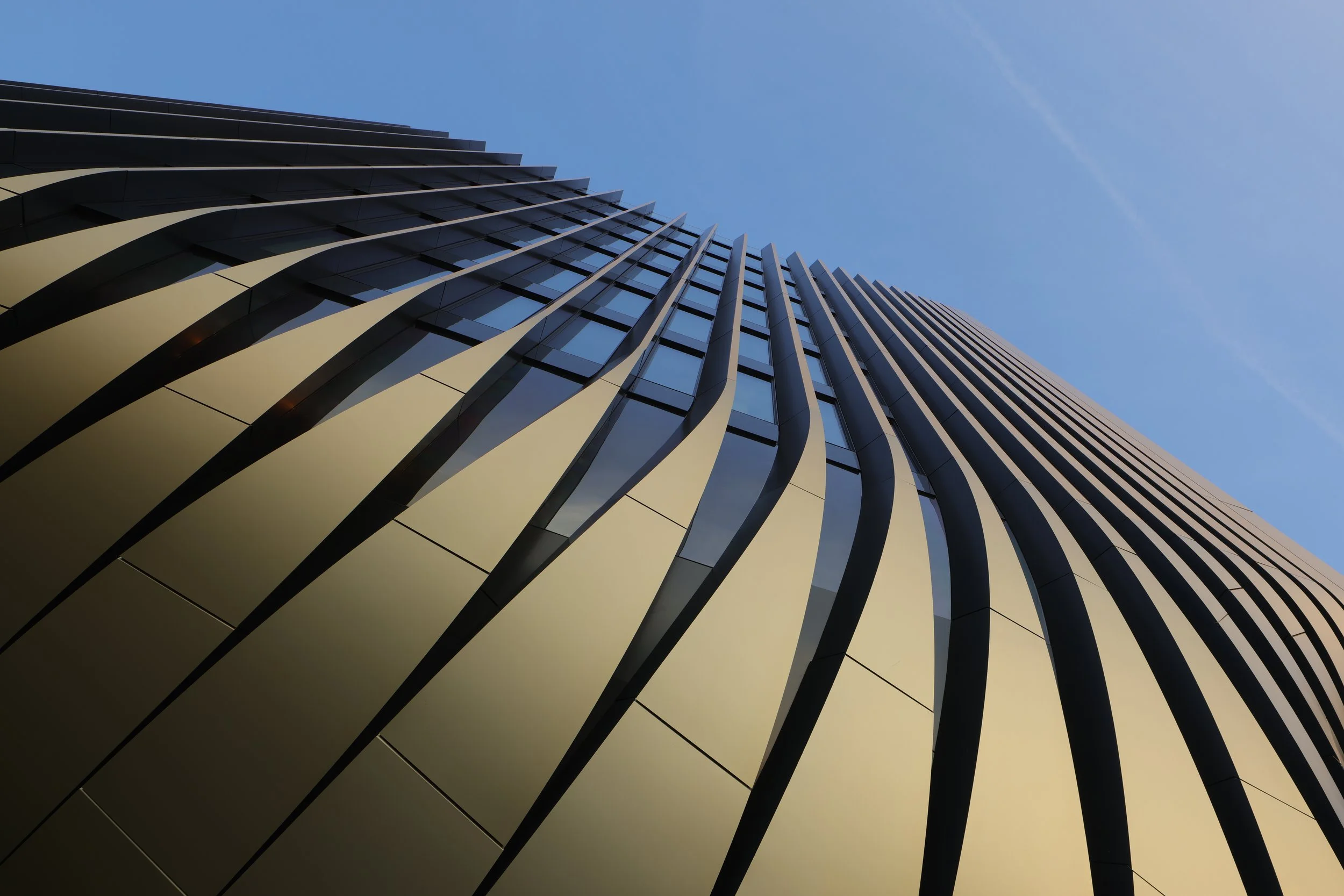 The exterior of a modern skyscraper with vertical golden panels and glass windows, viewed from the ground looking up, against a clear blue sky.