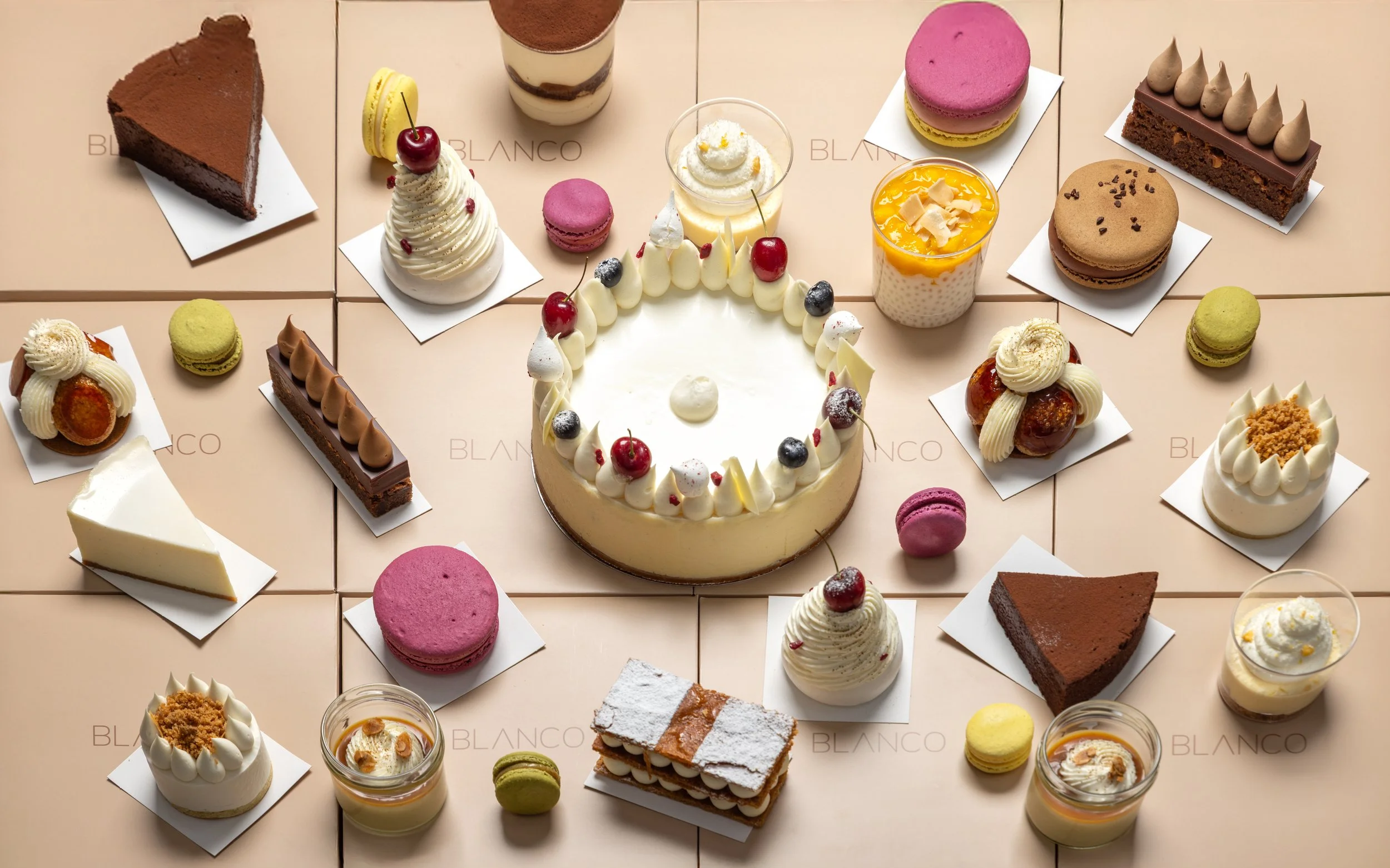 Assorted desserts including cake slices, macarons, tarts, and parfaits arranged on a tiled surface, with a central round cake decorated with cherries and cream.