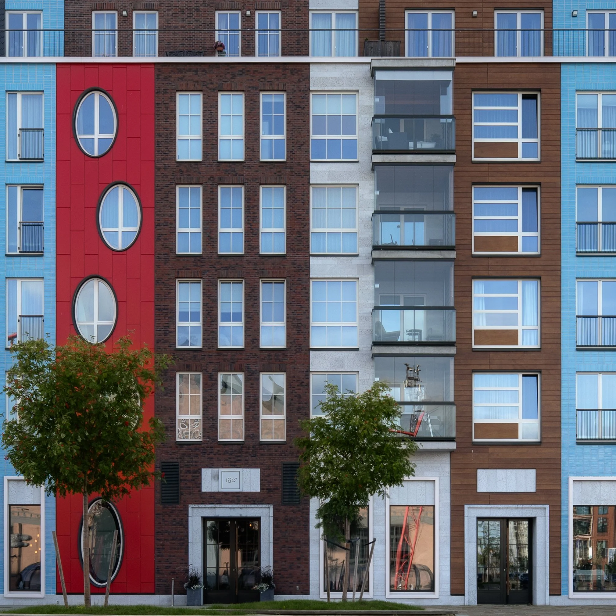 Front view of a modern multi-story apartment building with a colorful facade, featuring blue, brown, red, and gray sections, with large windows, some with balconies, and two small trees in front.