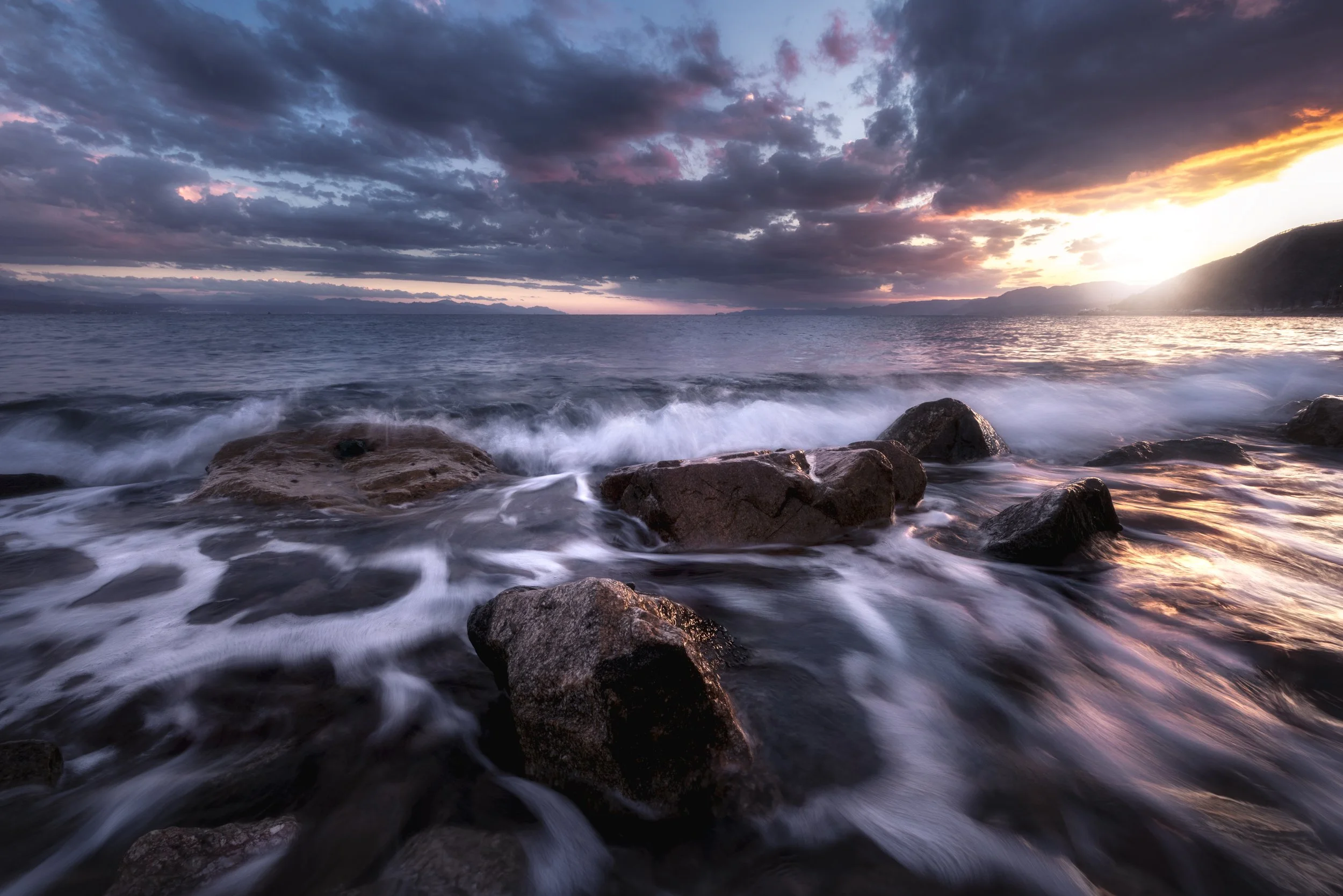 Sunset over a rocky shoreline with waves crashing against rocks and clouds in the sky.