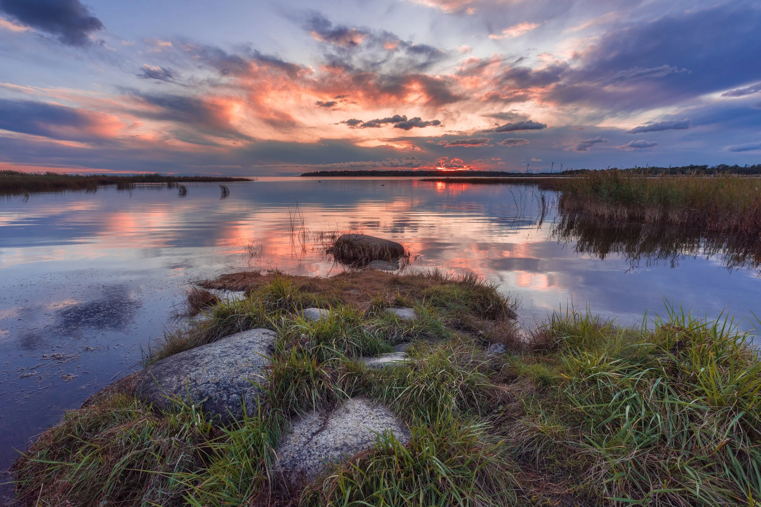 Sunset over a calm river with colorful clouds, grasses, and rocks along the riverbank.