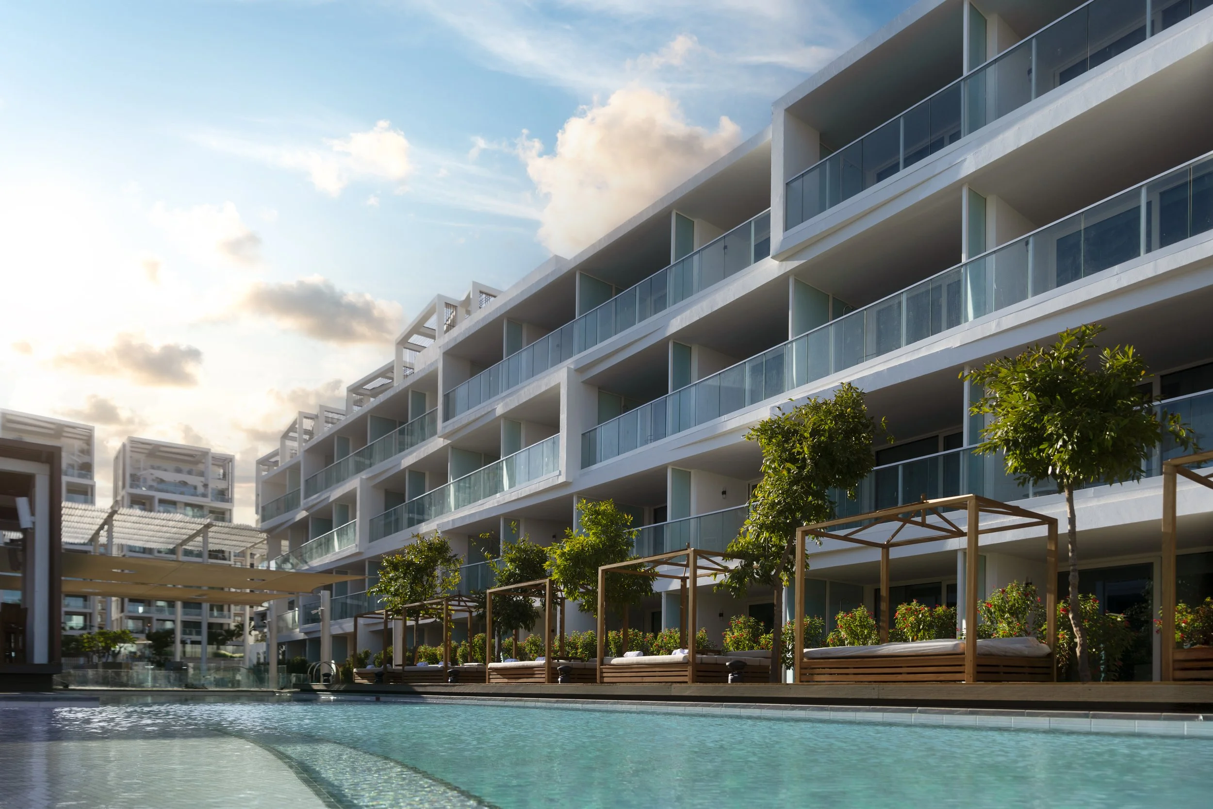 Luxury hotel with a swimming pool, outdoor seating, and landscaped trees under a partly cloudy sky.