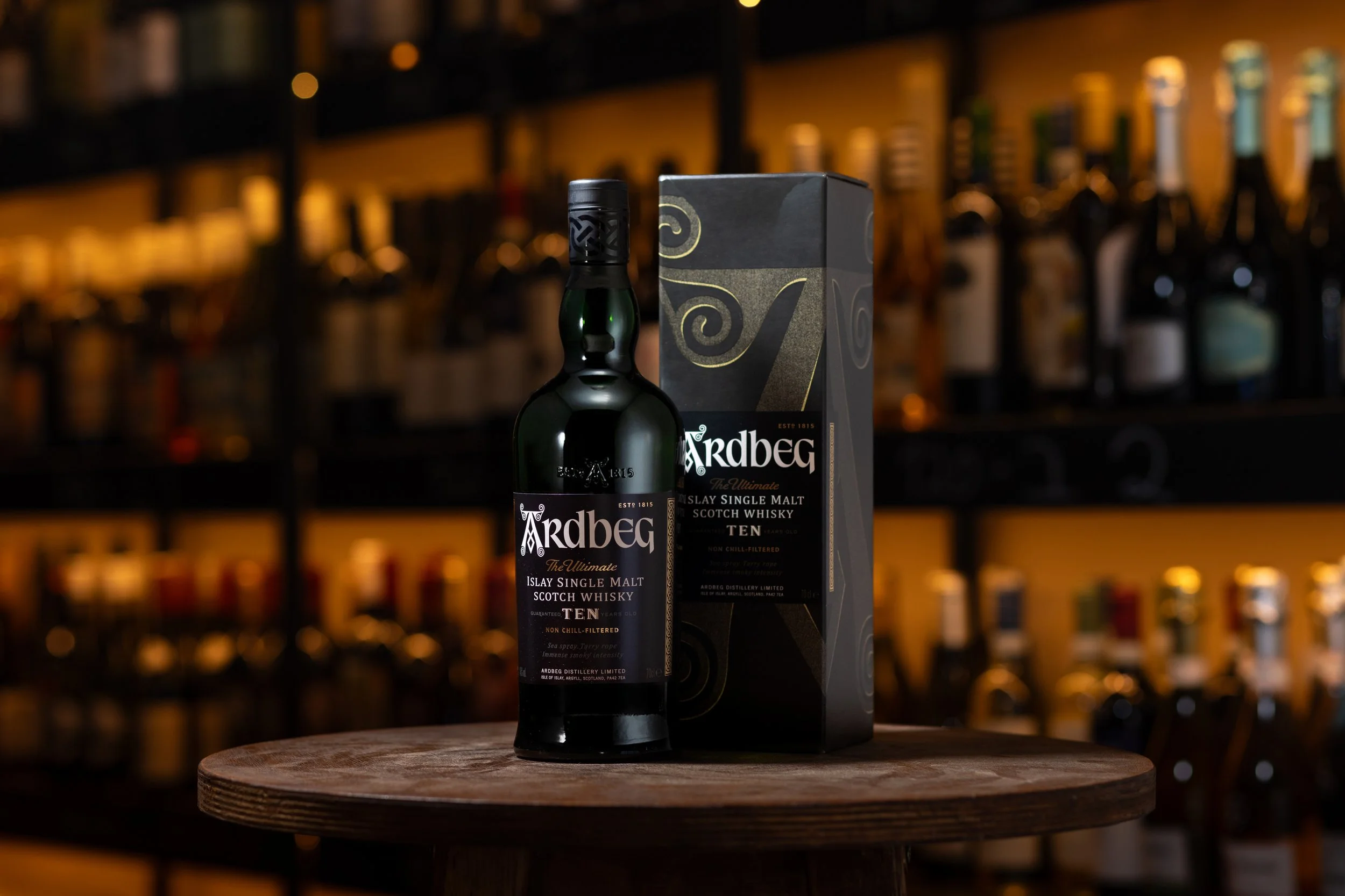 A bottle of Ardbeg Ten Year Old Islay Single Malt Scotch Whisky next to its box on a wooden table, with a blurred background of a liquor store shelf filled with various bottles.